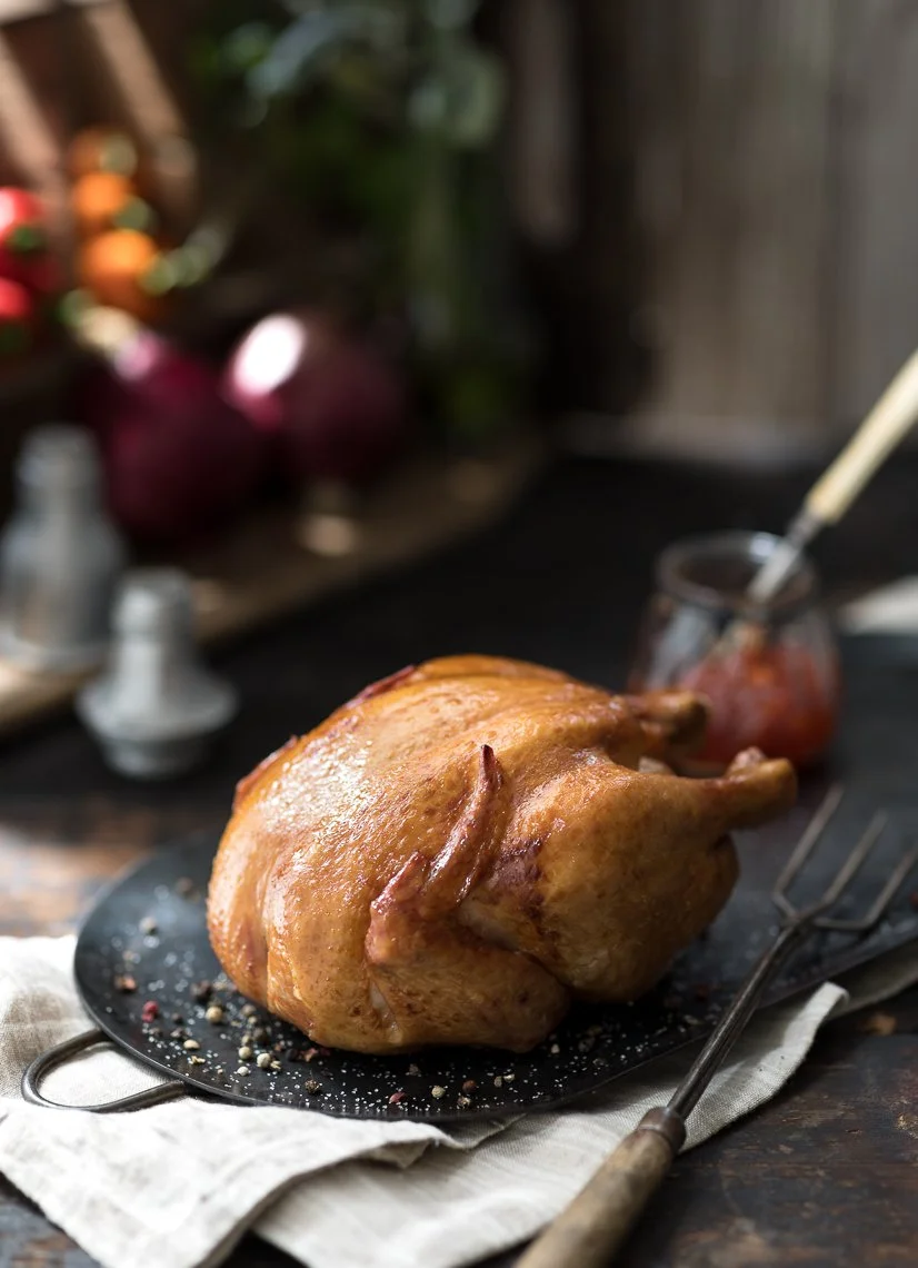 Roast chicken on a black serving plate with salt and pepper, placed on a wooden table with a white cloth underneath. Background includes a jar of tomato sauce and vegetables.