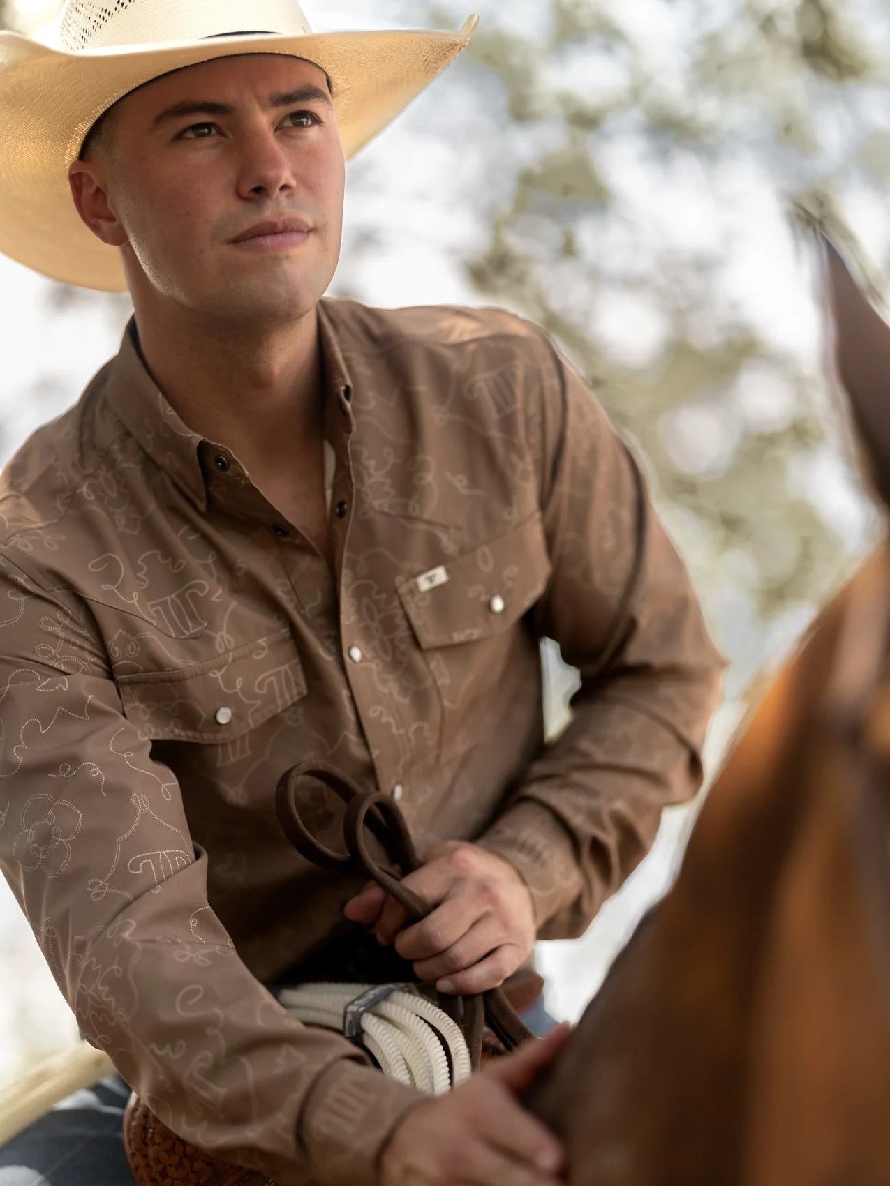 A young man wearing a cowboy hat and a brown western shirt is sitting on a horse, holding the reins, outdoors with blurred trees in the background.