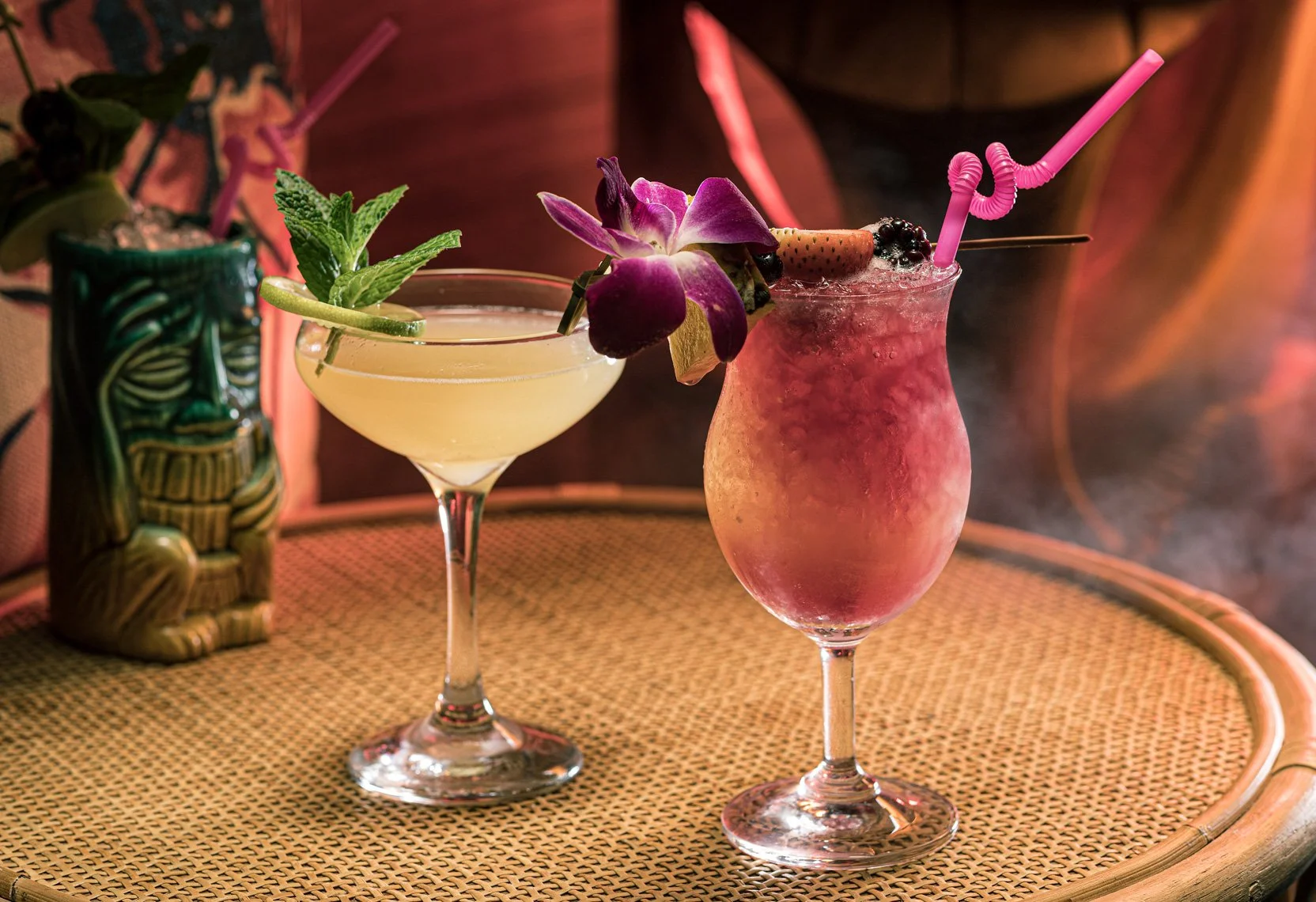 Two tropical cocktails on a rattan table, one with a lemon slice and mint garnish, the other with an orchid flower, strawberries, blackberries, and a pink straw.