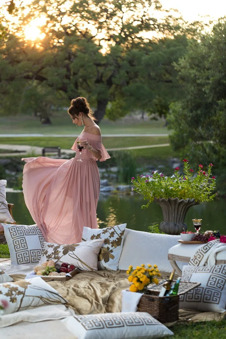A woman in a pink dress holding a glass of red wine standing on a blanket with pillows and food outdoors during sunset, with trees and a pond in the background.