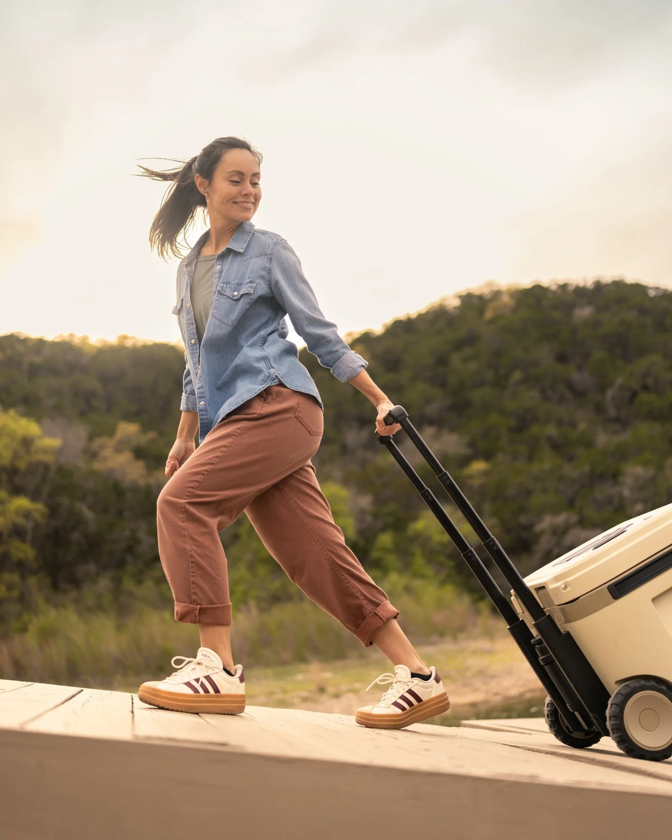 A woman with long hair, wearing a denim jacket, tan pants, and sneakers, is smiling and walking on a dock pulling a suitcase with a retractable handle, with a scenic background of trees and a cloudy sky.
