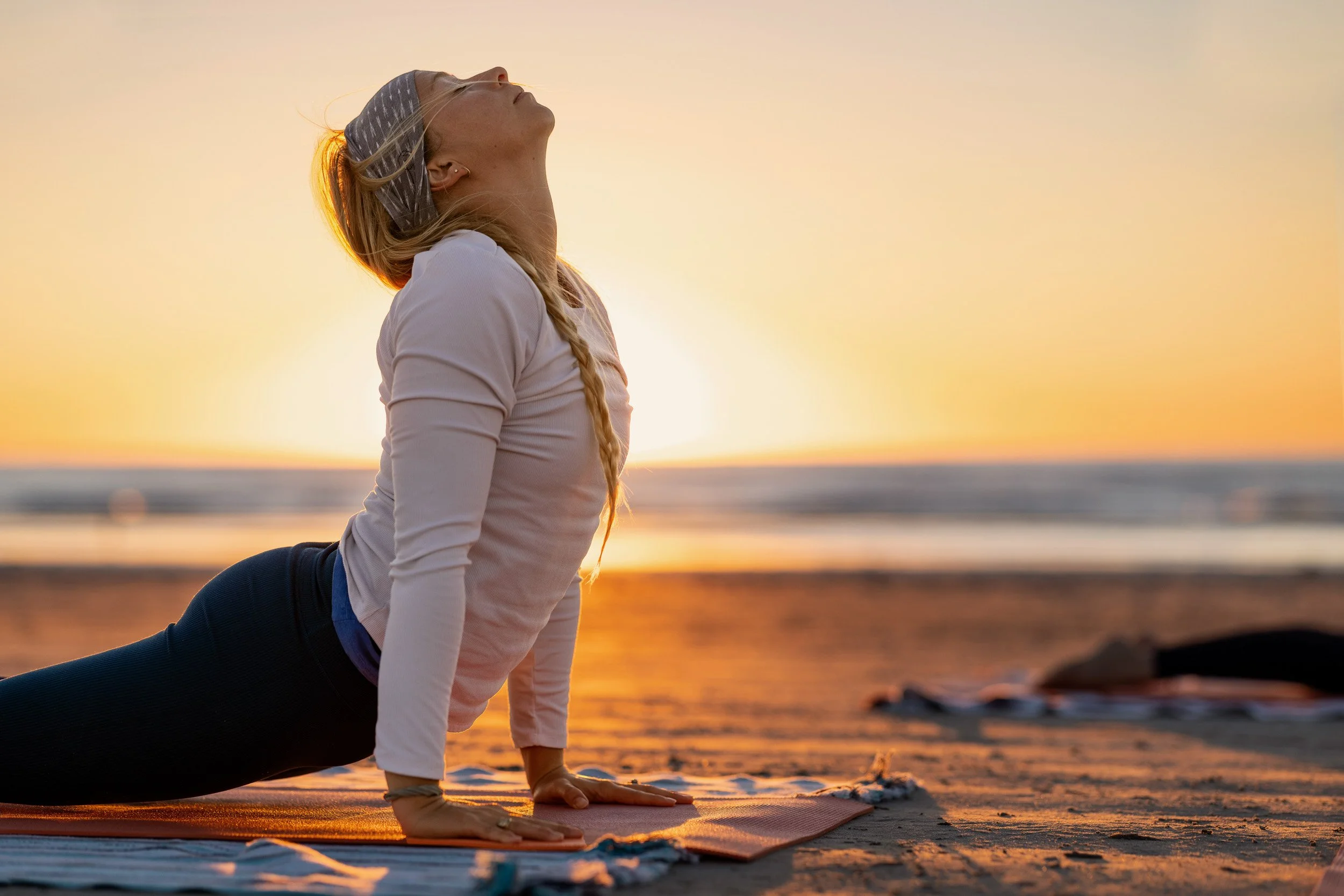 A woman practicing yoga in the cobra pose on a beach at sunset, with the ocean and sky in the background.