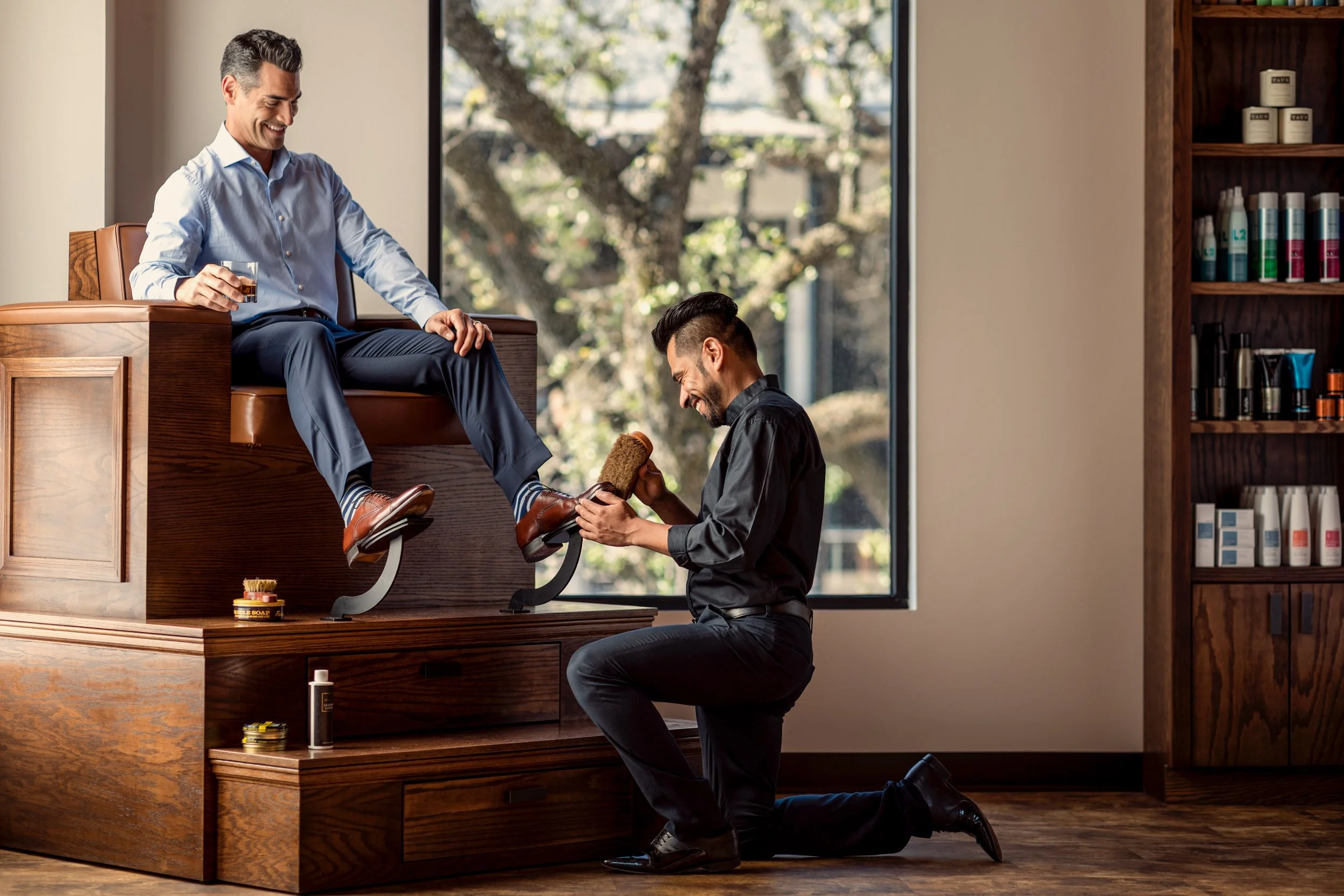 A man receives a pedicure at a salon, sitting on a wooden platform with his foot held by a professional man kneeling on the floor, both smiling and smiling. The man sitting has a glass of drink in his hand and the background shows a large window with