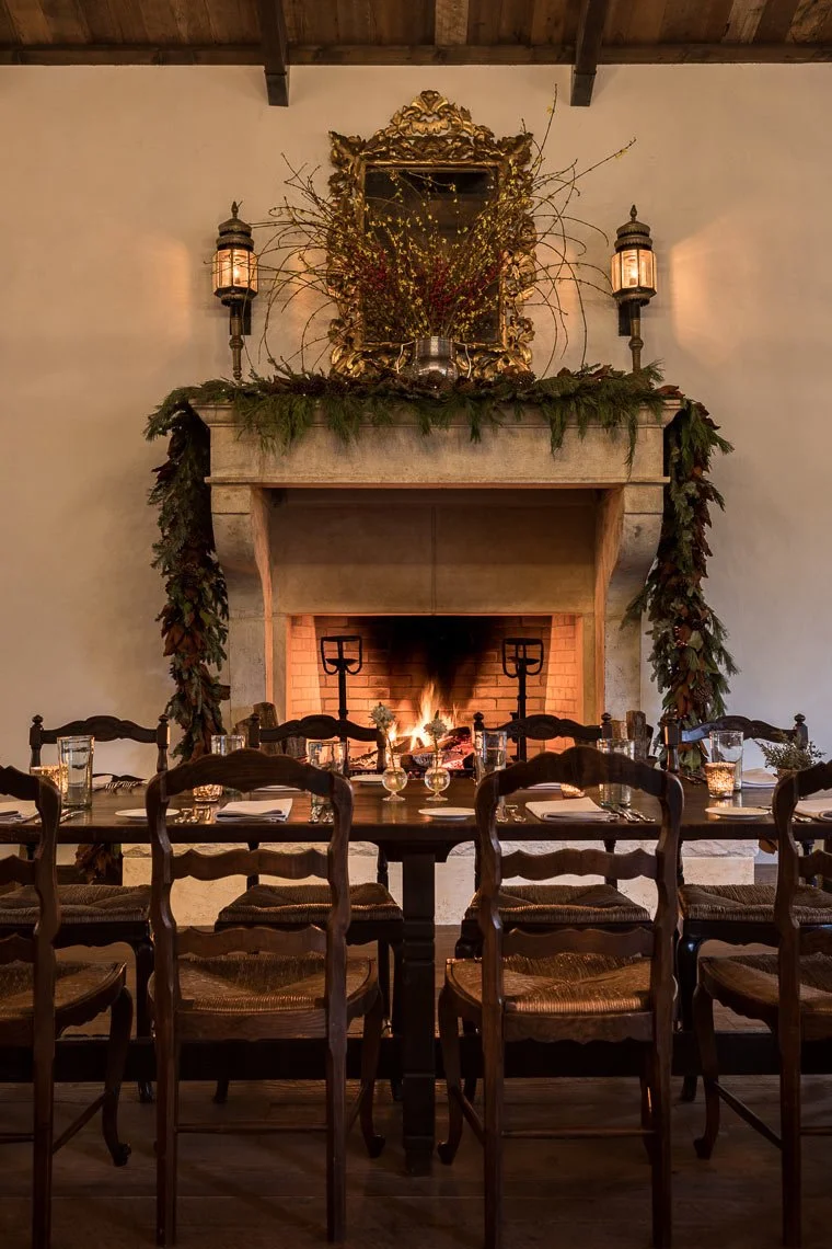 A cozy dining room featuring a large fireplace decorated with greenery, candles, and a mirror with ornate gold frame above it. A long dining table with chairs, glasses, and tableware is set in front of the fireplace, with a fire burning inside the he