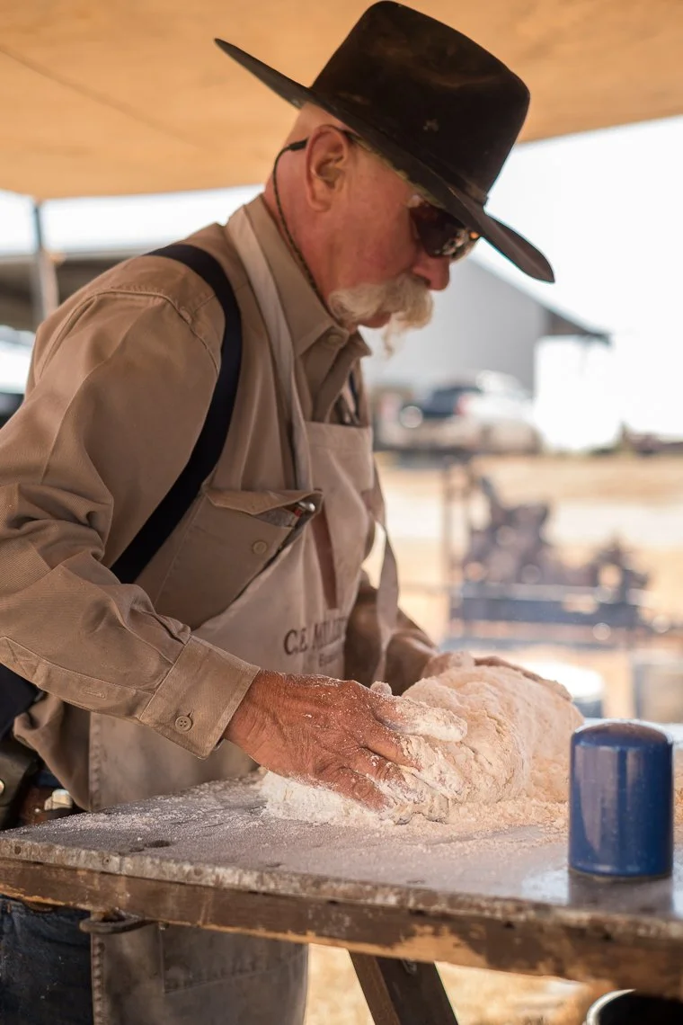 An elderly man with a gray beard, glasses, and a wide-brimmed hat kneading dough on a floured surface inside a rustic bakery or outdoor kitchen.