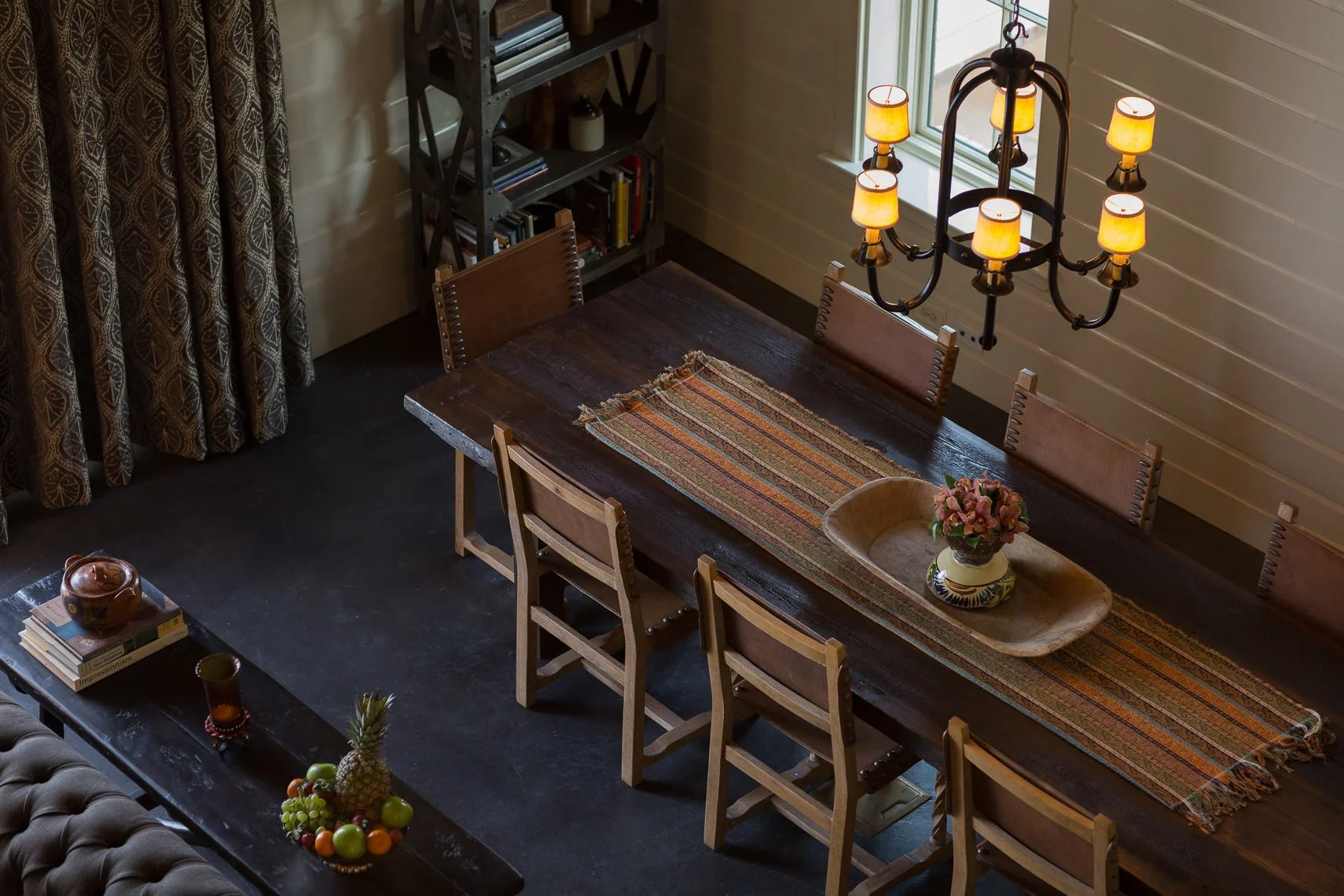 A dining room with a dark wooden table, six wooden chairs, and a colorful table runner. There is a vase with flowers on a tray on the table, and a fruit bowl with various fruits. Hanging above is a large black chandelier with multiple yellow lamps. I