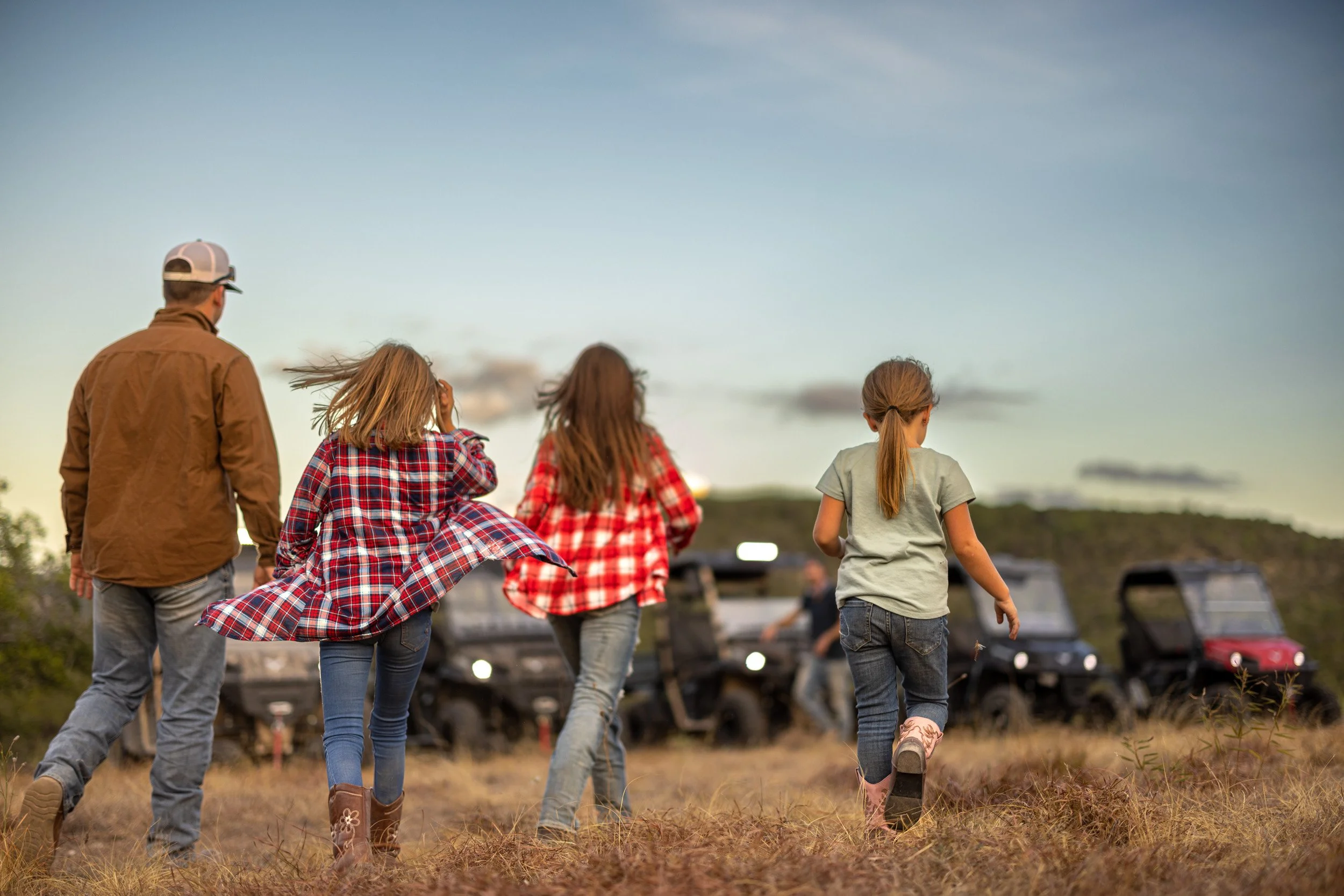 Five people, three children and two adults, are walking on a grassy field during dusk or early evening, with a cloudy sky overhead and cars parked in the background.