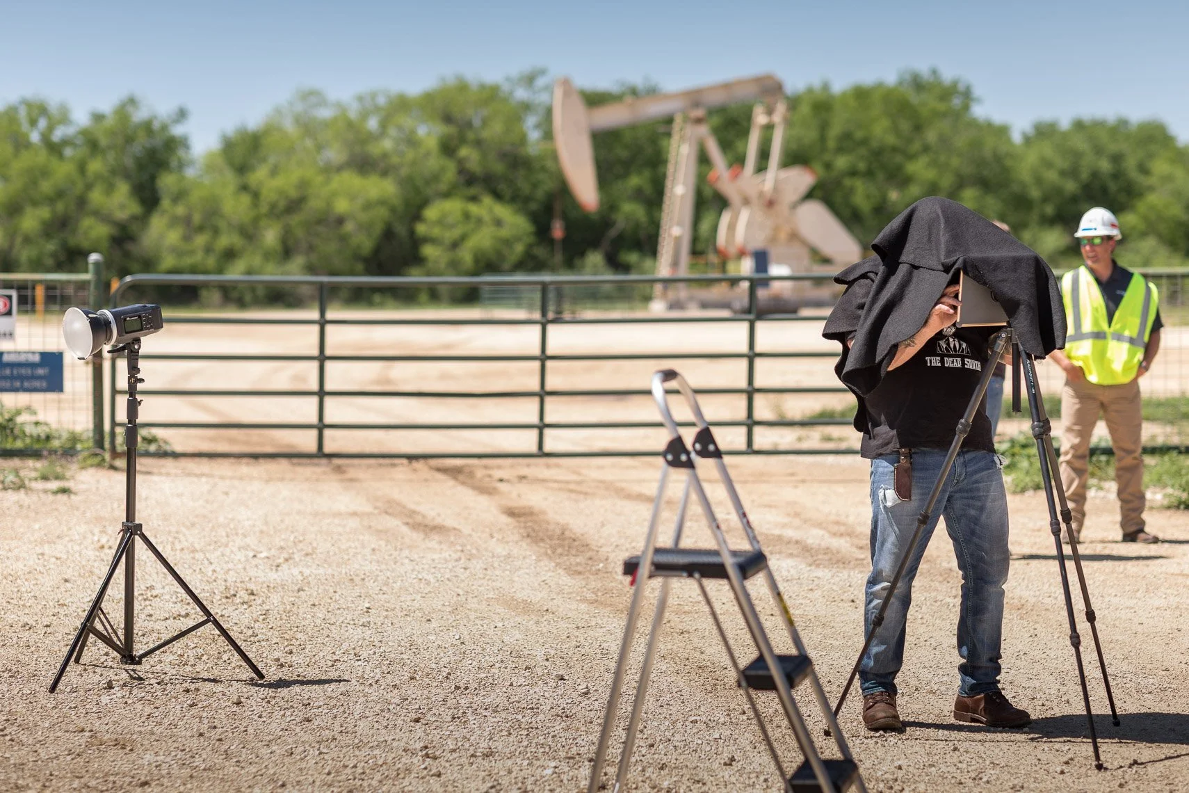 Photographers set up camera equipment at a construction site with a large excavator in the background and a man standing by a black fence.