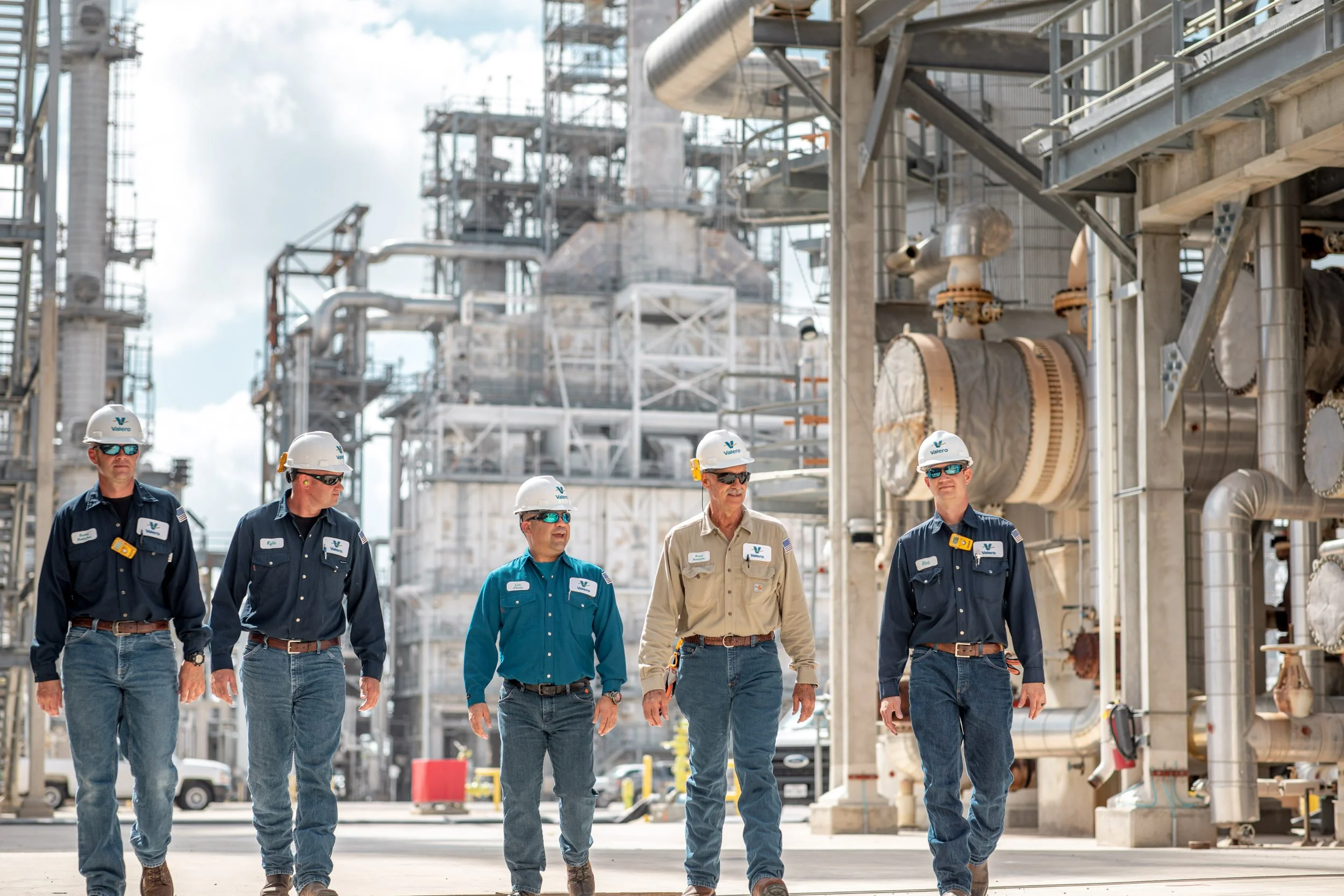 A group of six workers walking through an industrial plant wearing hard hats, safety glasses, and work uniforms, with large machinery and metal structures in the background.