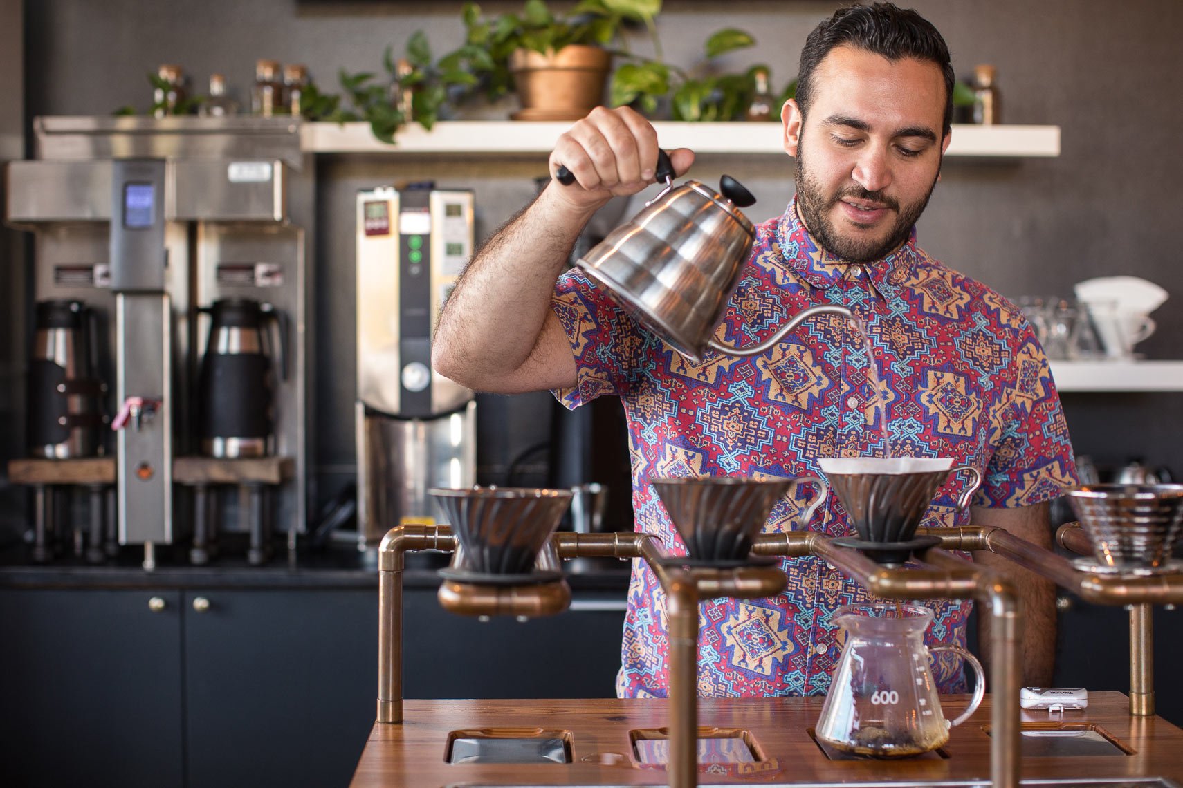 A man in a colorful patterned shirt pouring hot water from a gooseneck kettle into a coffee dripper, surrounded by coffee brewing equipment and accessories on a wooden table in a coffee shop.