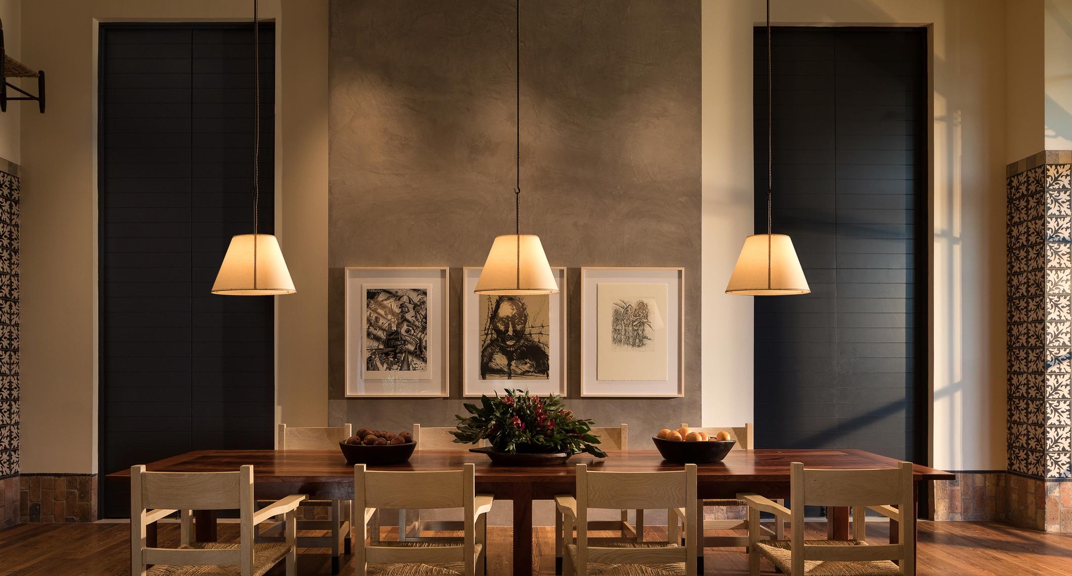 Interior of a dining area with a wooden table, four chairs, three framed artworks on a gray wall, a bowl of fruit, and hanging pendant lights.