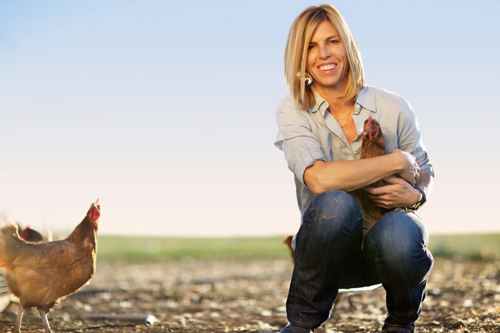 A woman crouching outdoors in a farm field, holding a chicken, with other chickens nearby, under a clear blue sky.