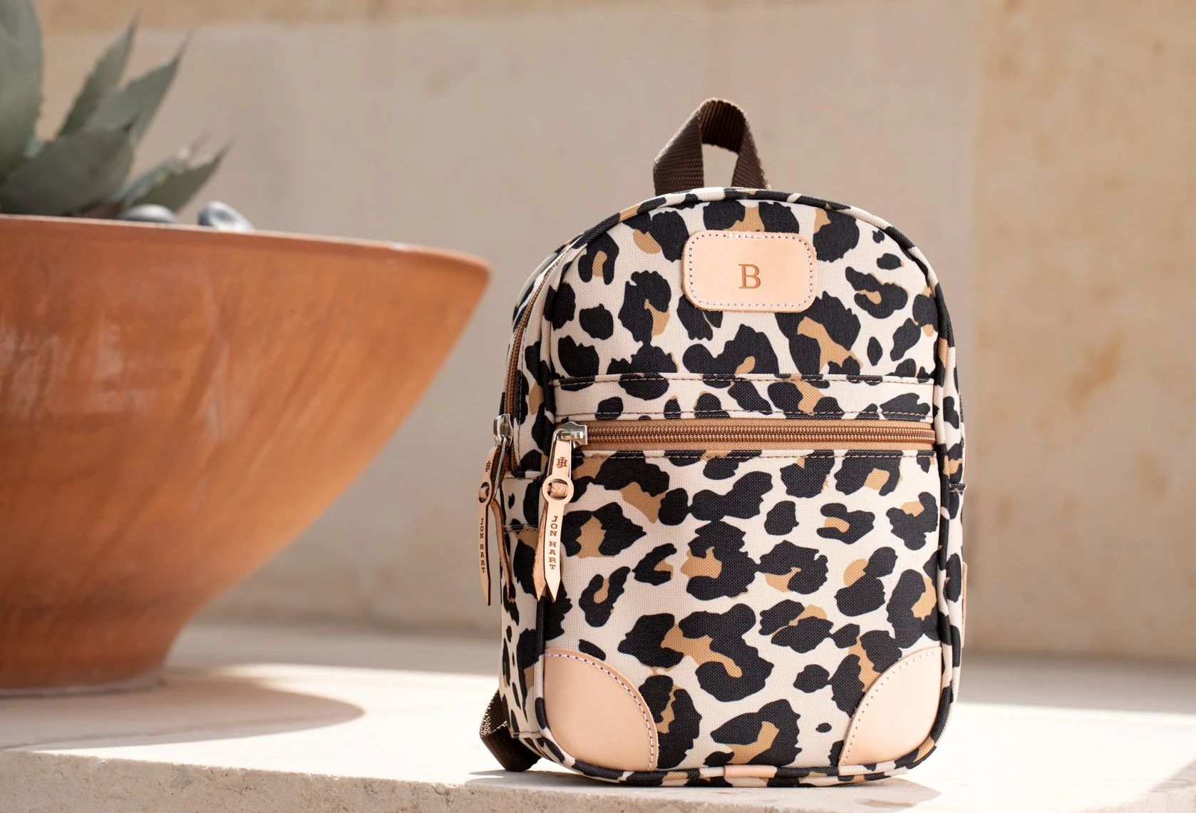 Leopard print backpack with beige accents, zippers, and a monogrammed label, placed on a white surface next to a large terracotta pot with a plant.