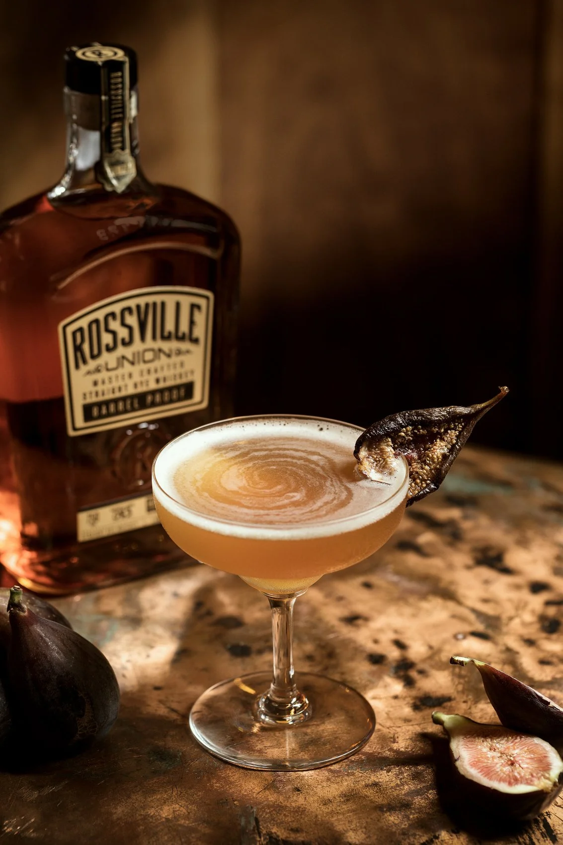 A cocktail with a pear wedge garnish on the rim, placed on a wooden surface with ripe pears around, and a bottle of Rossville Union bourbon in the background.
