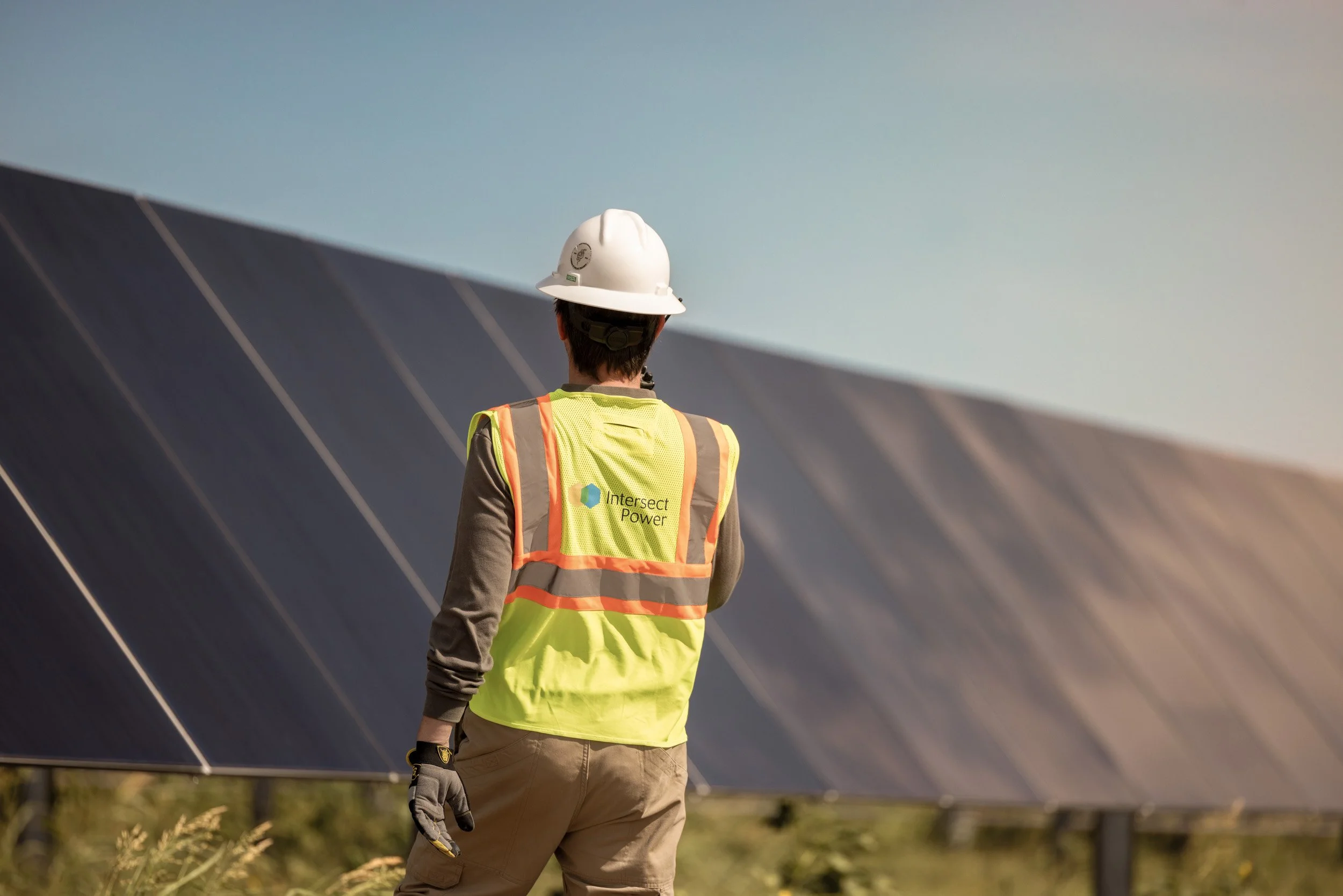 A person wearing a white hard hat and a yellow safety vest with 'Intersect Power' logo is inspecting solar panels outdoors during daytime.