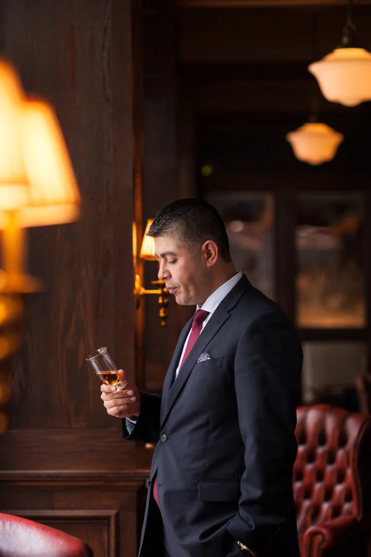 A man in a dark suit holding a glass of whiskey inside a warmly lit, wood-paneled room with red leather chairs and hanging pendant lights.