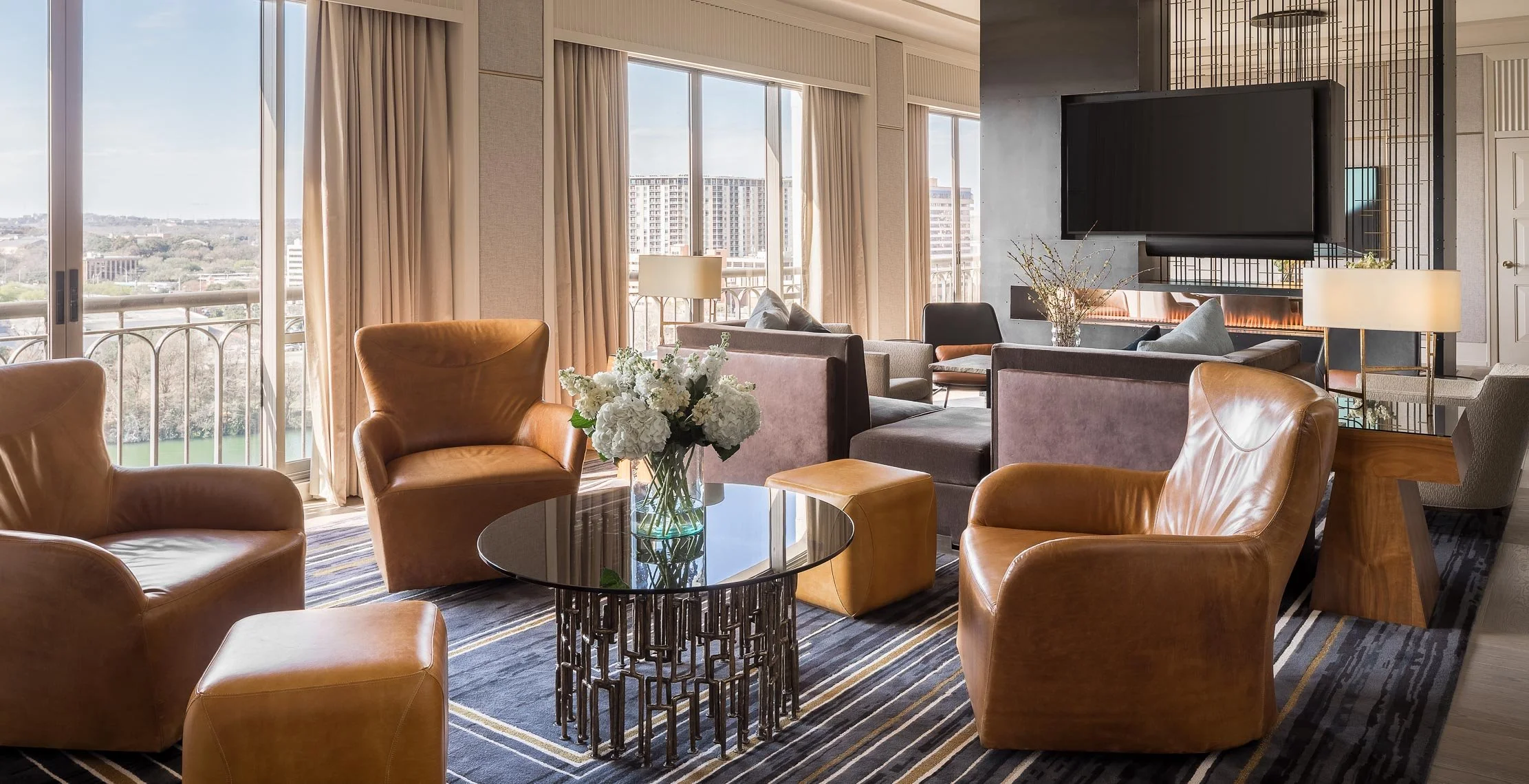 A modern hotel lounge with large floor-to-ceiling windows, beige curtains, and city views. The seating area includes leather armchairs, upholstered sofas, and a glass coffee table with a floral arrangement. A flat-screen TV is mounted on a wall above