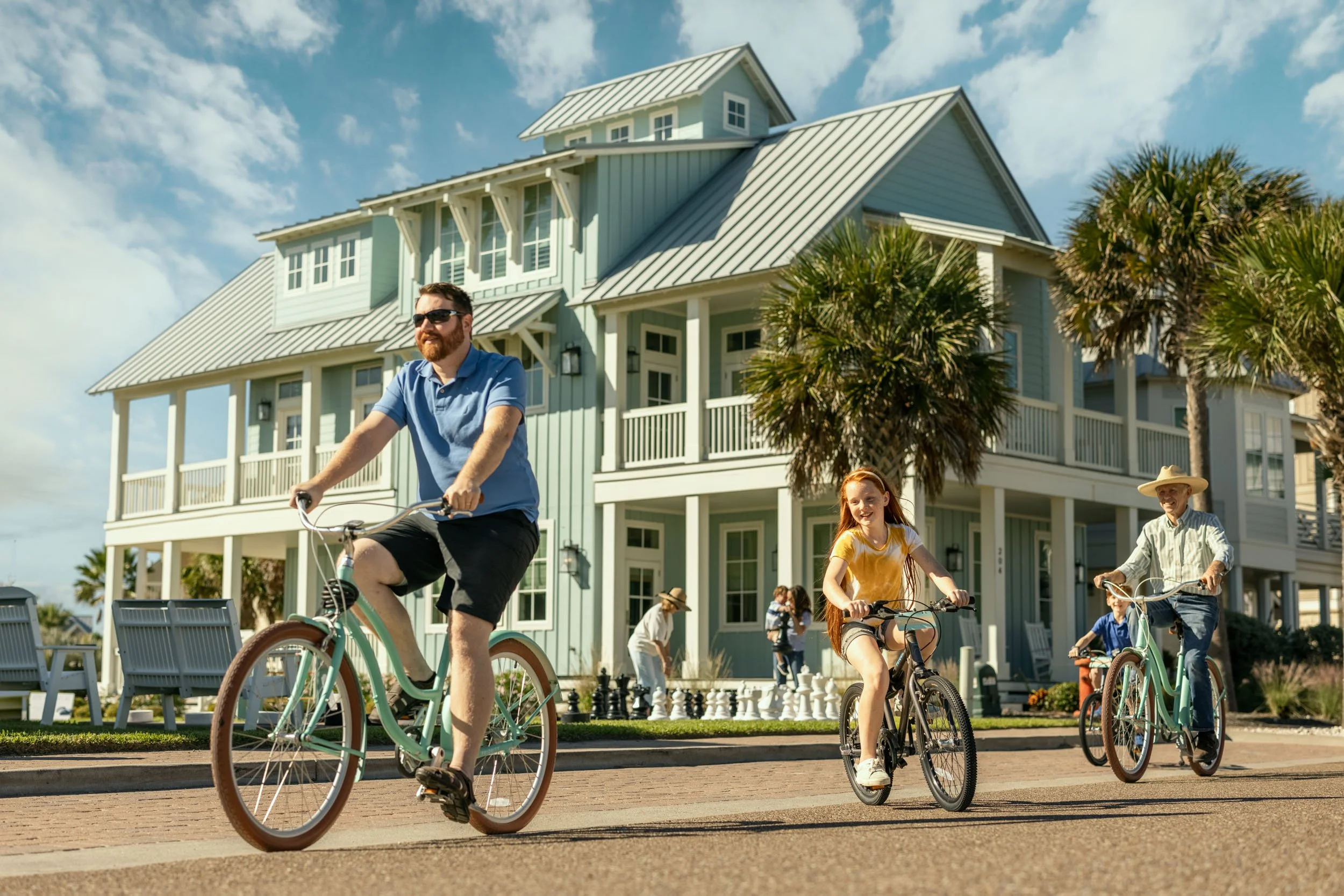 People riding bicycles in a neighborhood with large multi-story houses and palm trees.
