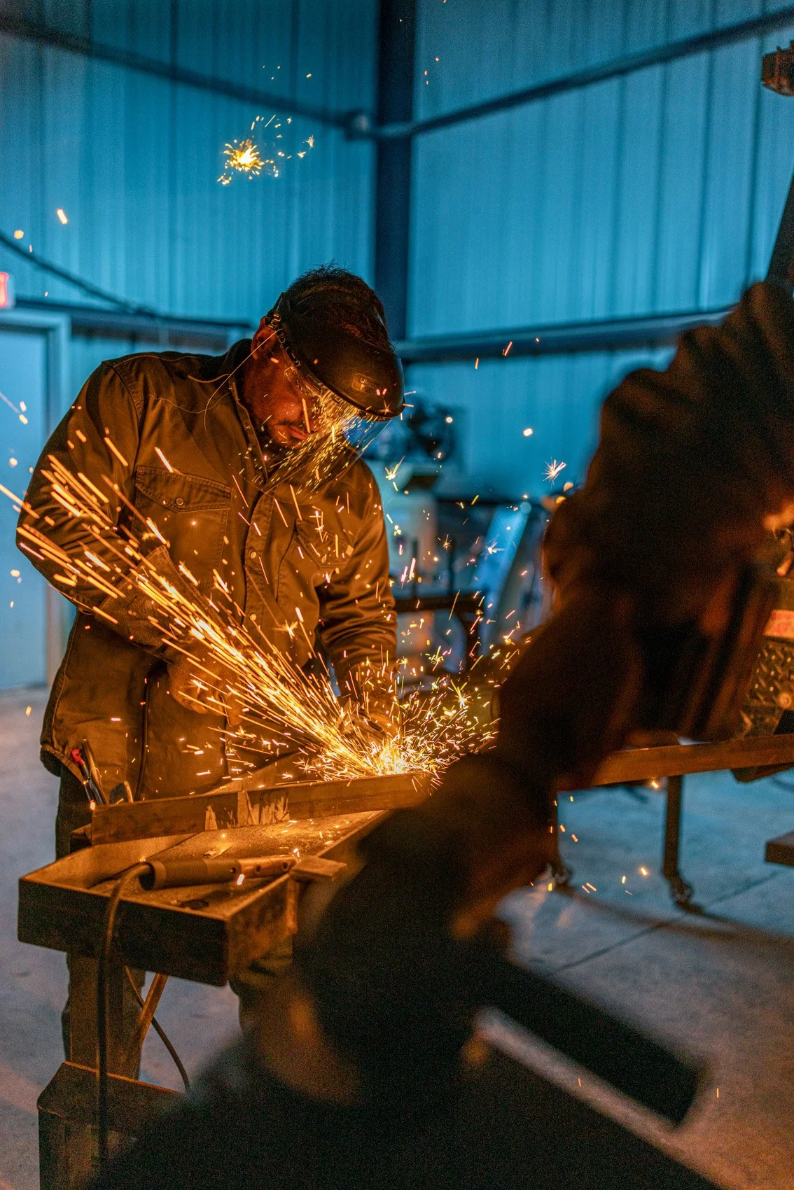 A person welding metal indoors, wearing a protective helmet and gloves, with sparks flying from the welding process.