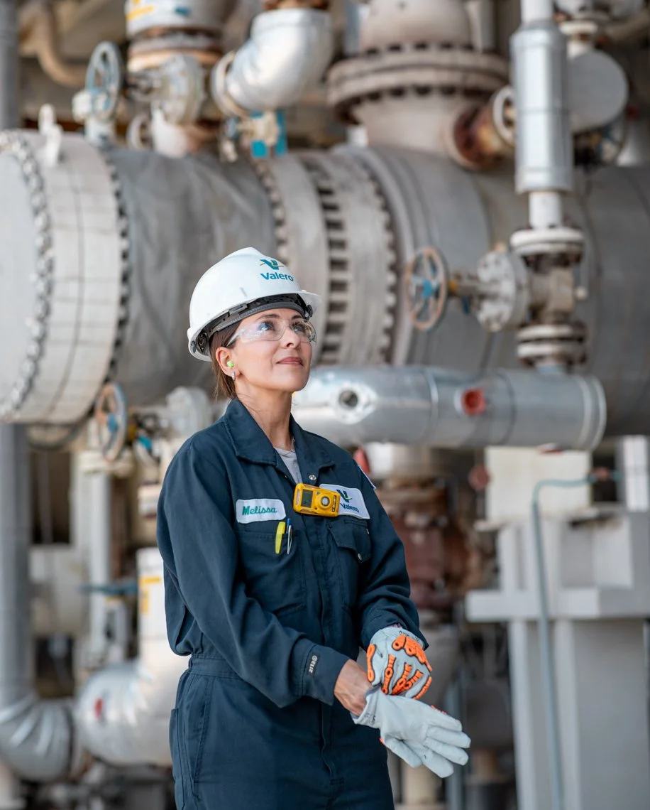 A woman wearing safety glasses, a white hard hat with the Valero logo, and a navy work jumpsuit stands in an industrial setting with large pipes and machinery behind her. She is holding work gloves and looking slightly upward.