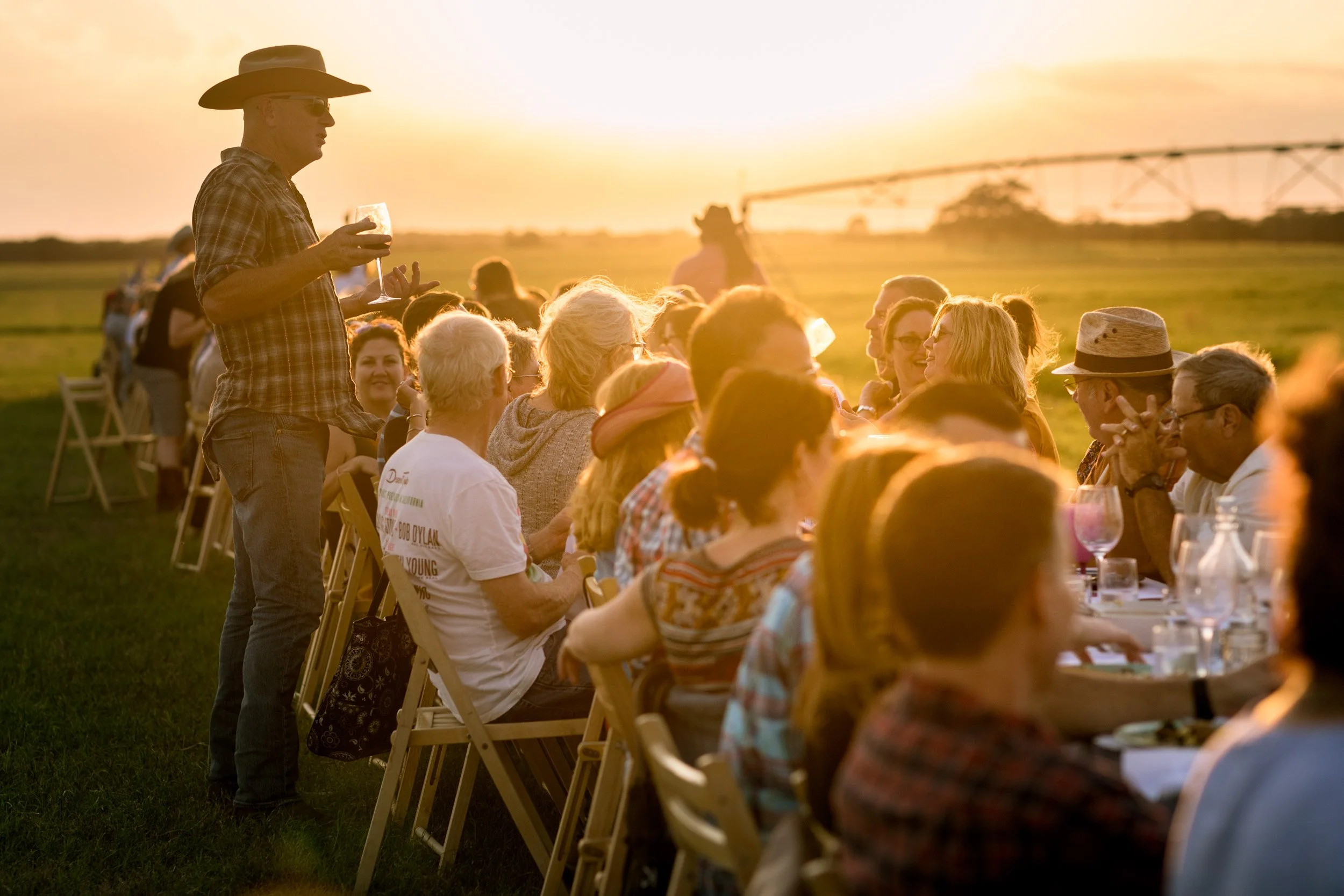 People gathered around a long table outdoors at sunset, enjoying a meal and drinks. A man in a cowboy hat and sunglasses stands and talks to the group, holding a glass of wine.