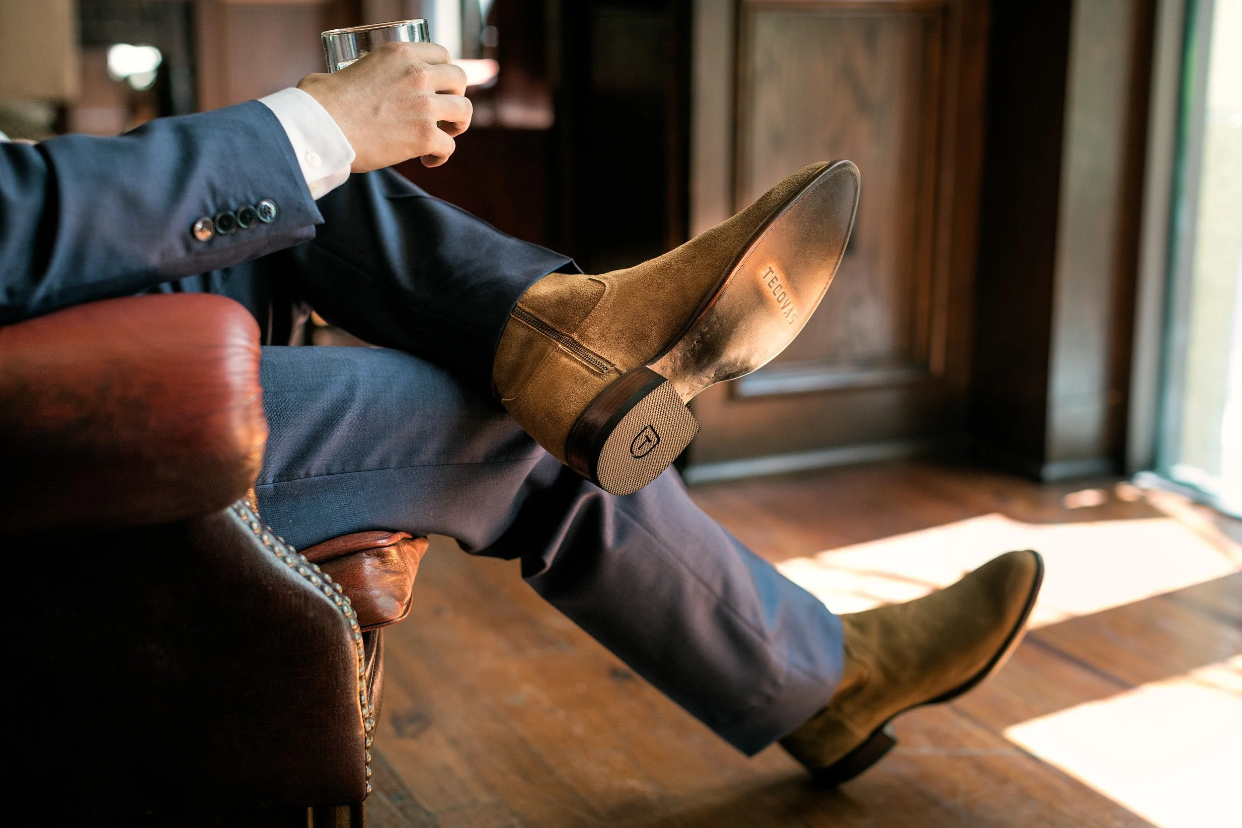 A person wearing a dark suit and brown suede boots sitting on a leather chair inside a room with wooden floors, holding a clear glass of water.