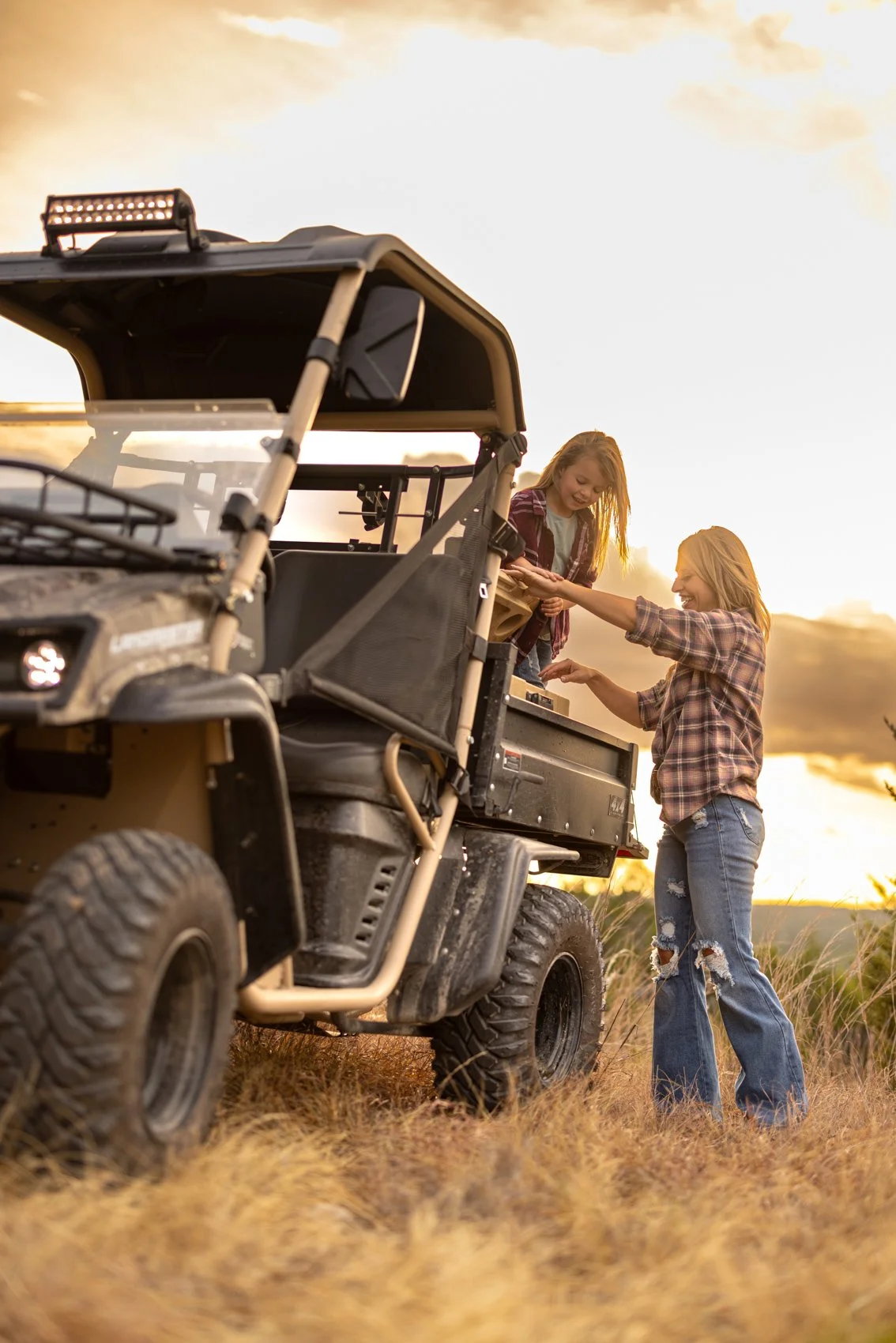 A woman and a girl sharing a moment of joy next to an off-road utility vehicle in a field during sunset.