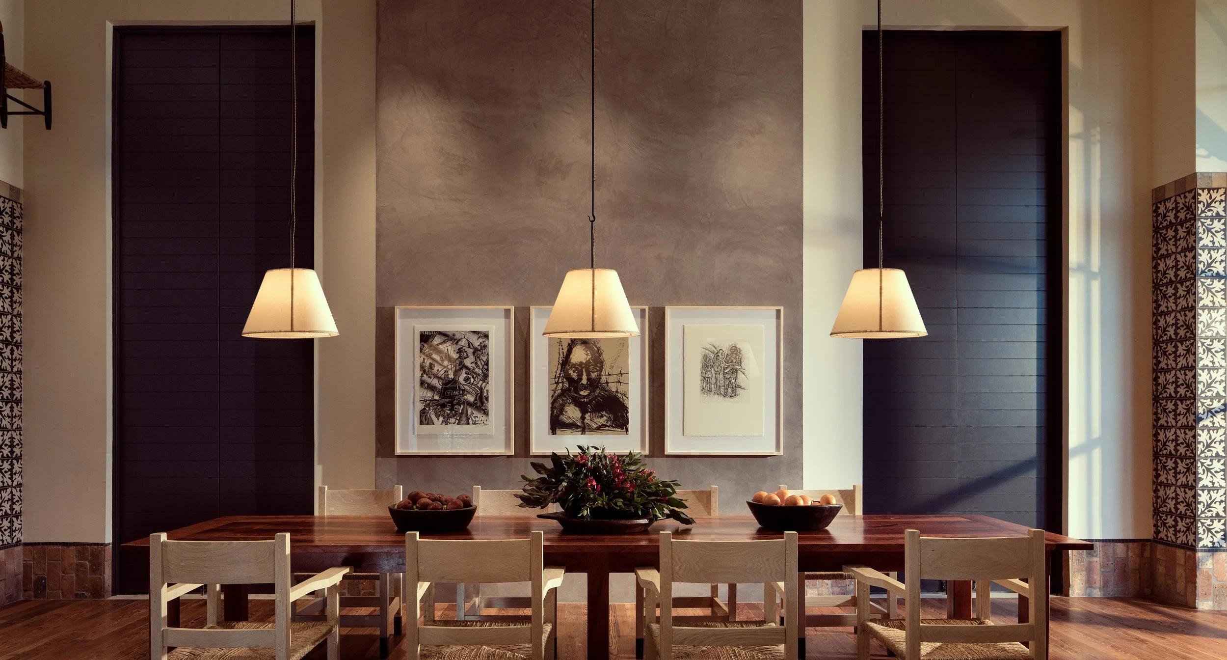 Modern dining room with three framed black and white art pieces on a beige wall, three hanging pendant lights, a wooden dining table, chairs, and bowls of fruit and flowers as centerpieces.