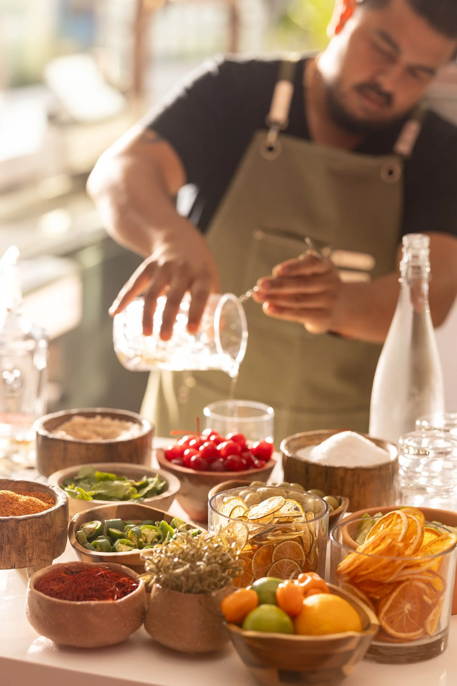 A man prepares food with various bowls of colorful fruits, vegetables, spices, and condiments on a table. The setting appears warm and well-lit, likely a kitchen or dining area.