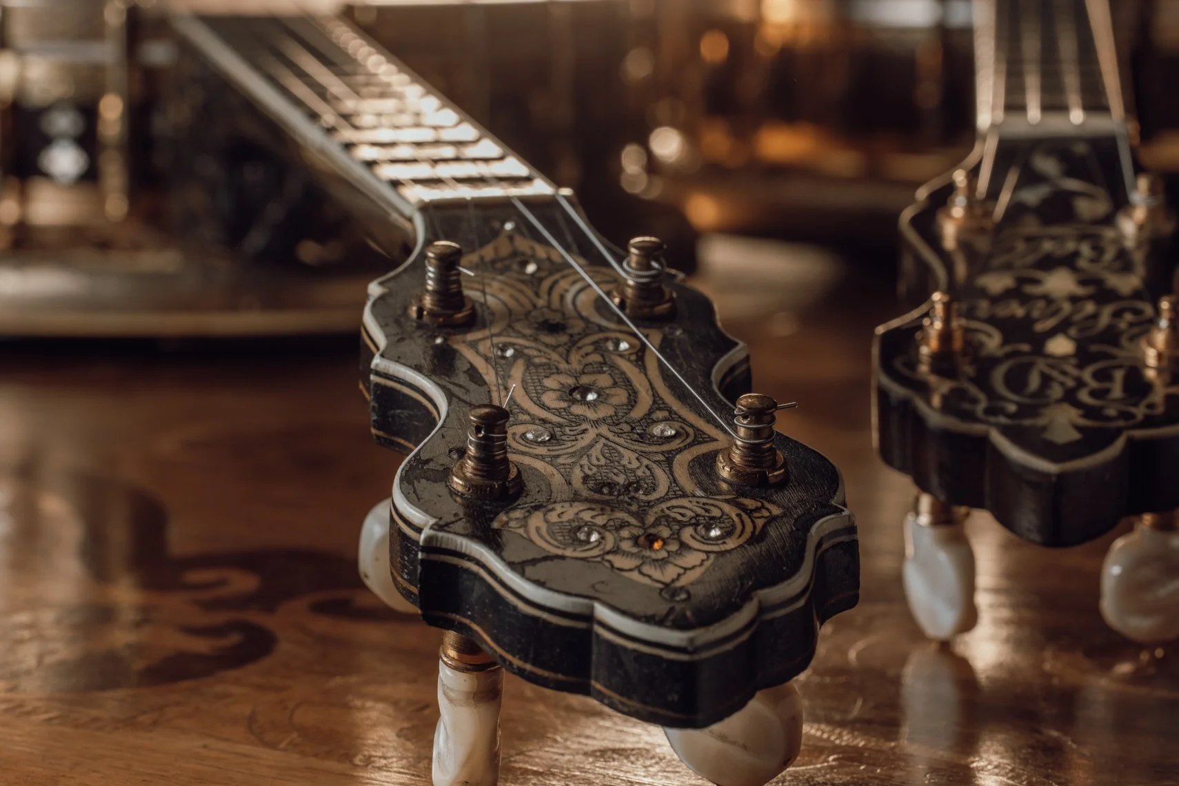 Close-up of an ornate, decorative guitar headstock with intricate black and cream design, gold tuning pegs, and white tuning machine knobs, placed on a wooden surface.