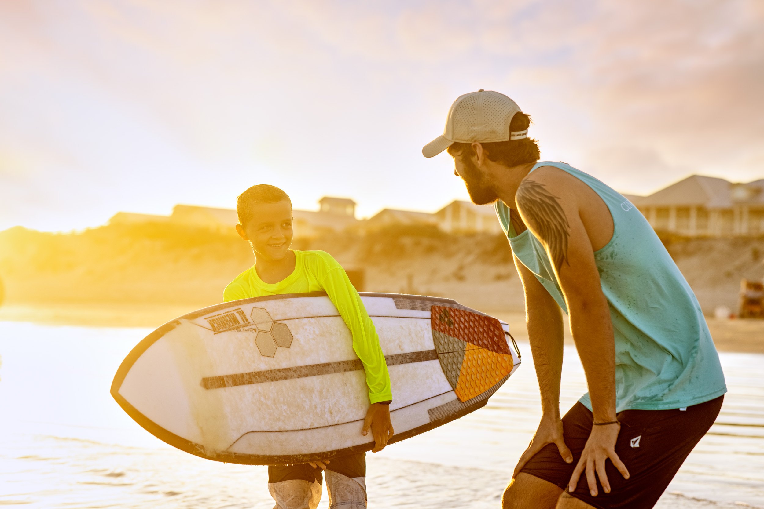 A man and a young boy on a beach at sunset, with the boy holding a surfboard and the man assisting him.