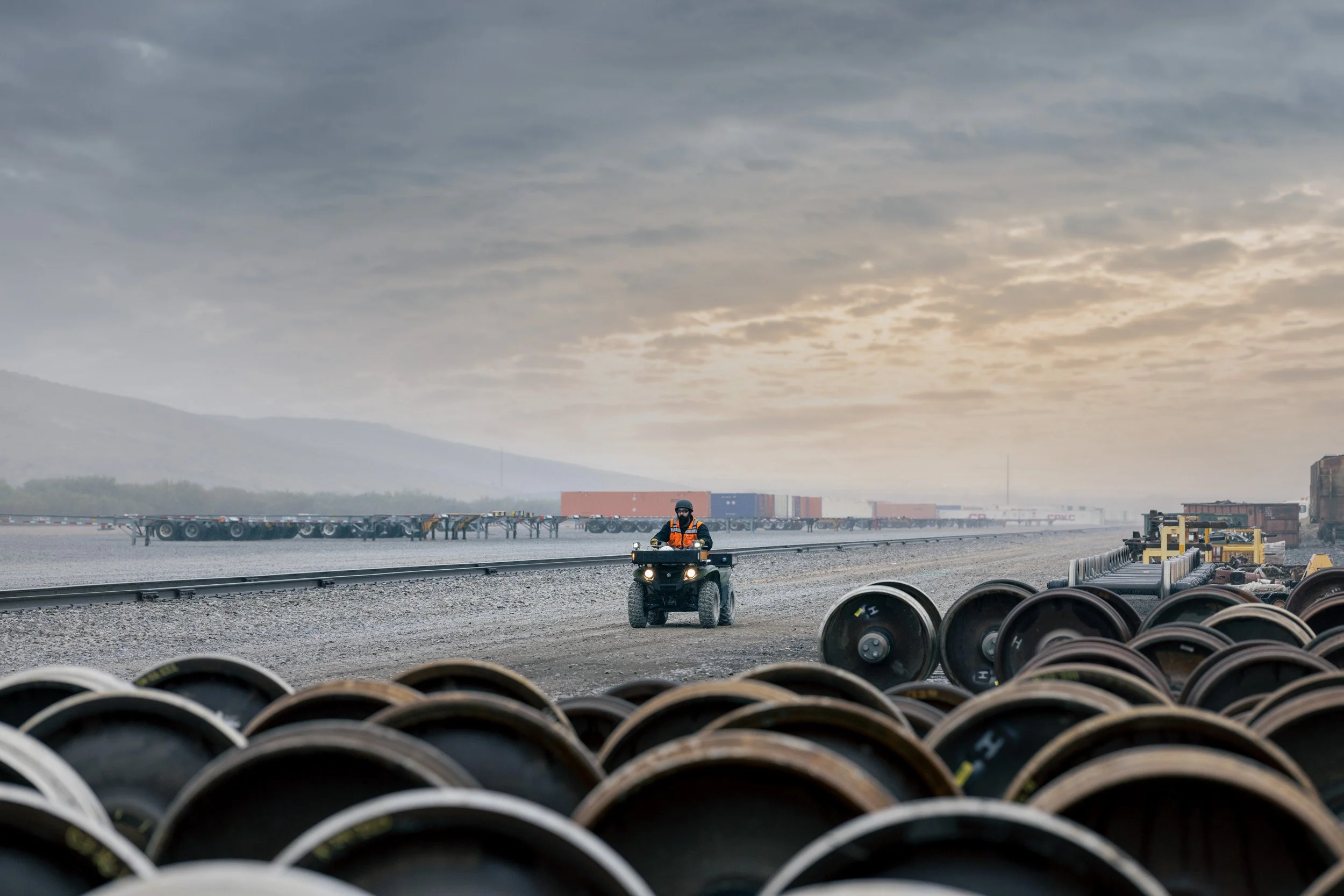 Industrial scene with tire stacks in the foreground, a person on an ATV in the middle ground, and shipping containers with mountains and cloudy sky in the background.