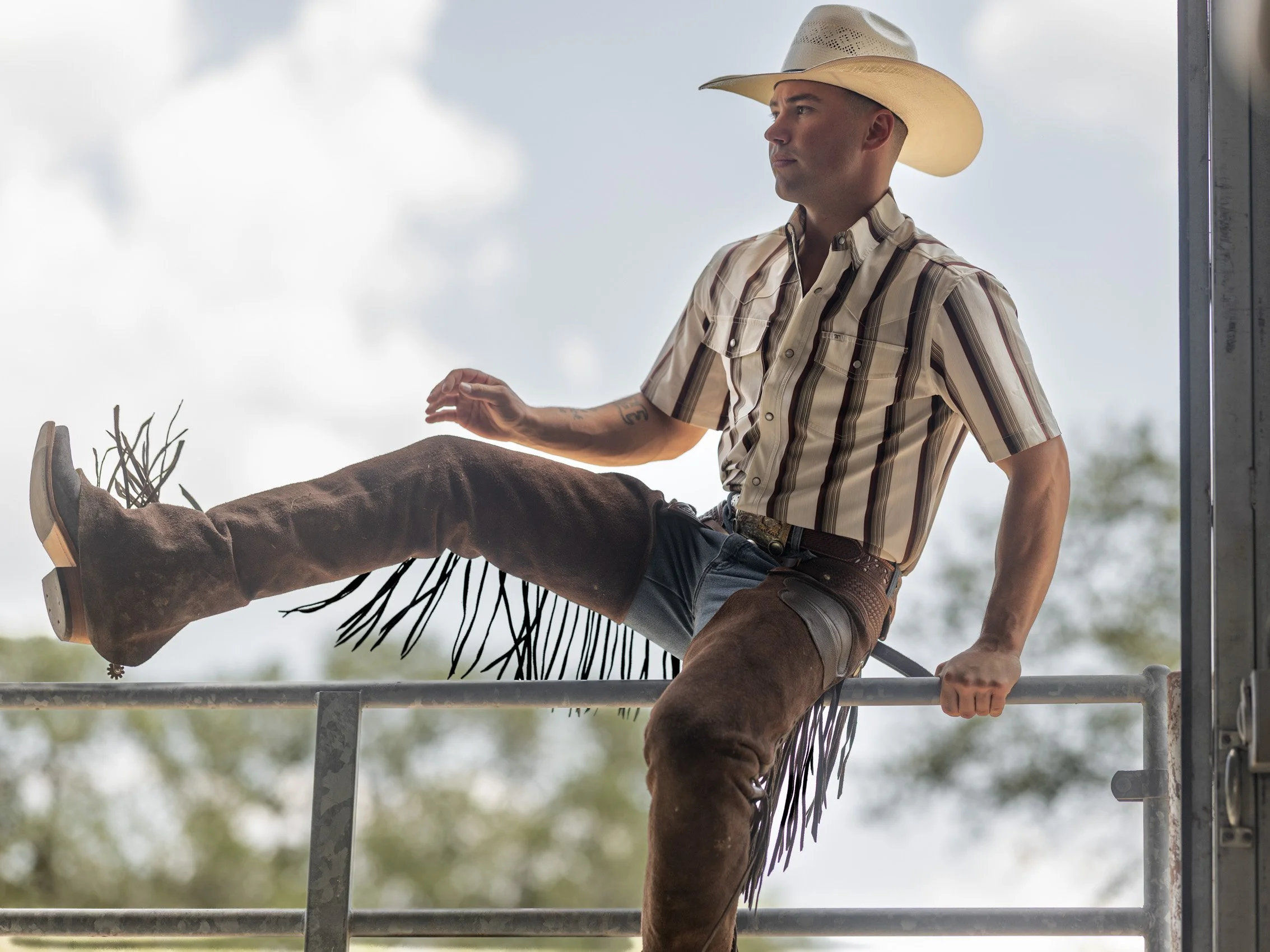 A man dressed as a cowboy, wearing a white cowboy hat and a plaid shirt, is sitting on a metal railing with one leg outstretched and the other bent at the knee. He is outdoors with a cloudy sky and trees in the background.