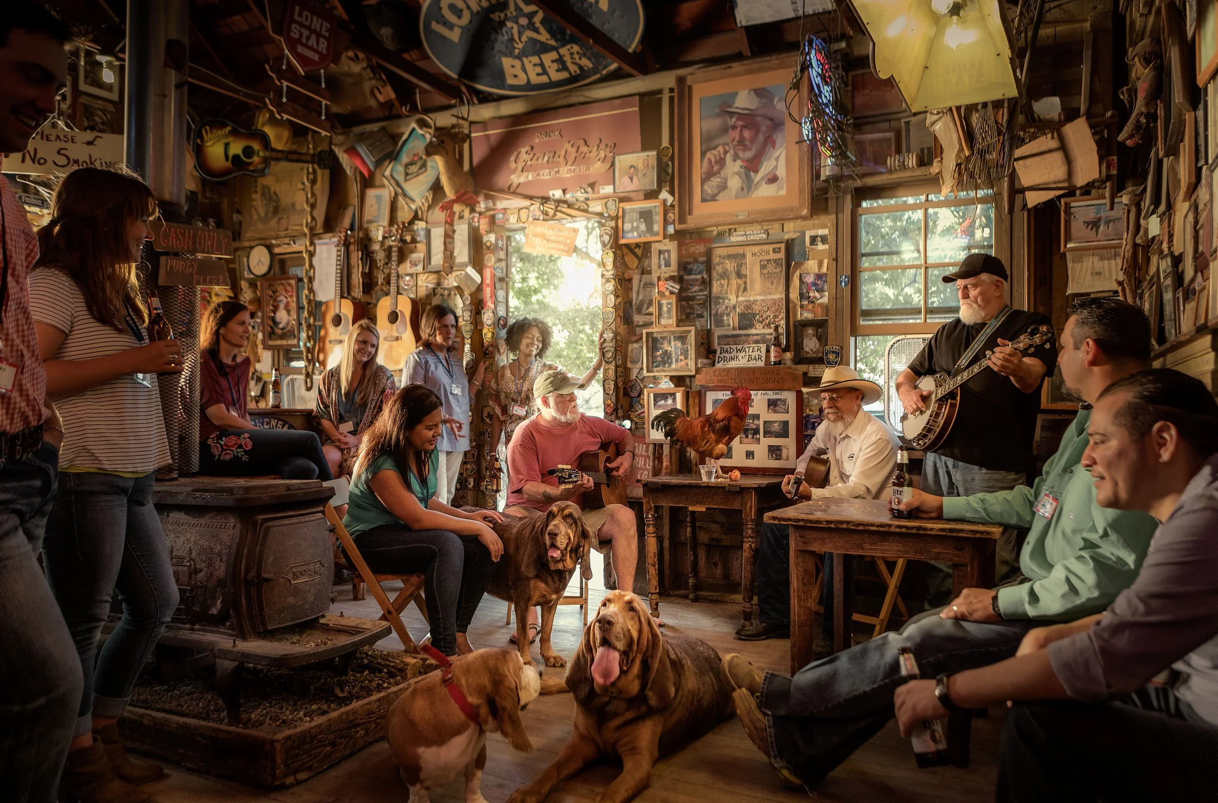 People gathered in a rustic, decorated bar, some sitting and some standing, while a man plays guitar. Dogs are on the floor, and the bar is adorned with guitars, photos, and signs.