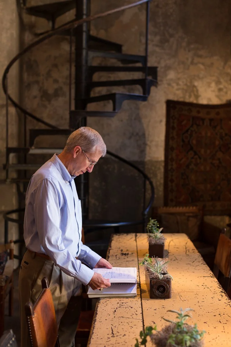 An older man with glasses, wearing a light blue shirt and khaki pants, stands at a large wooden table reading a notebook in a rustic room with exposed brick walls and a spiral staircase.