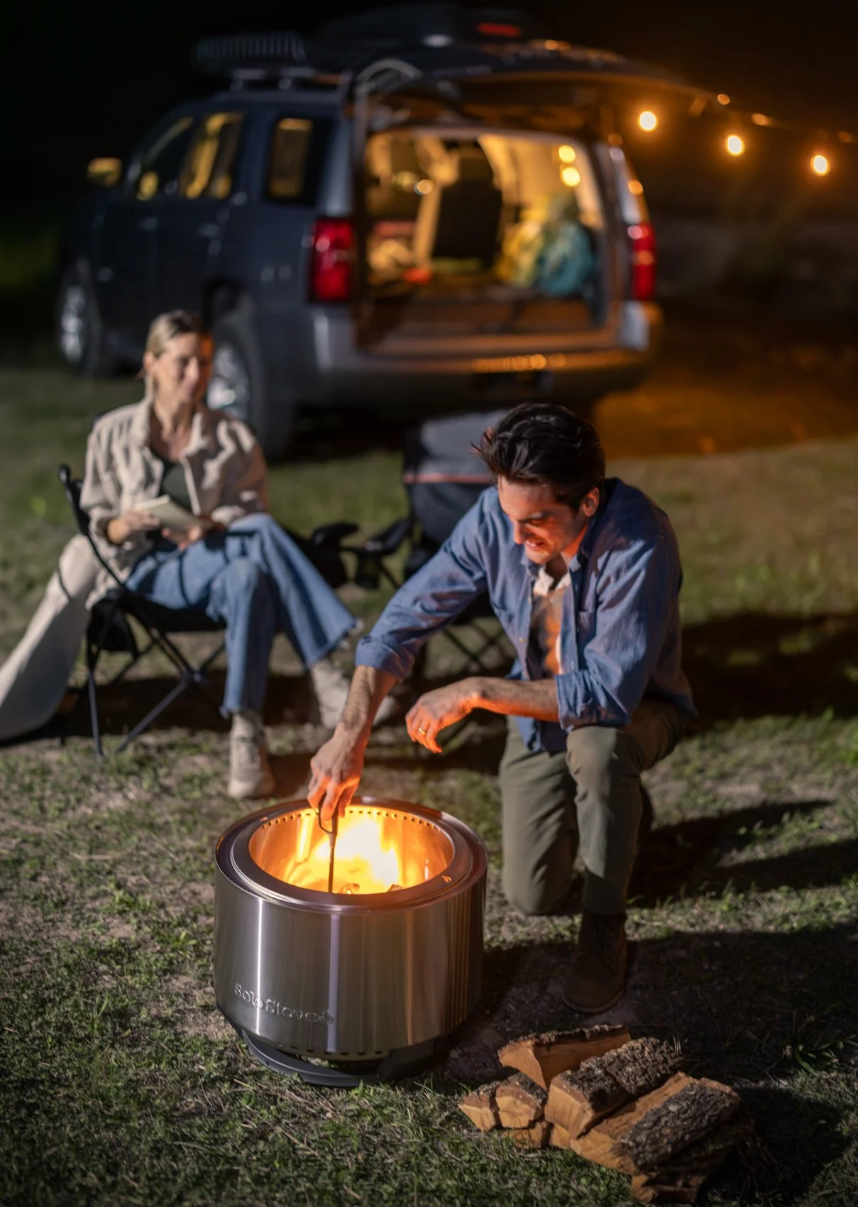 People sitting by a vehicle at night with open trunk, one person adding wood to a portable fire pit, and firewood on the ground nearby.