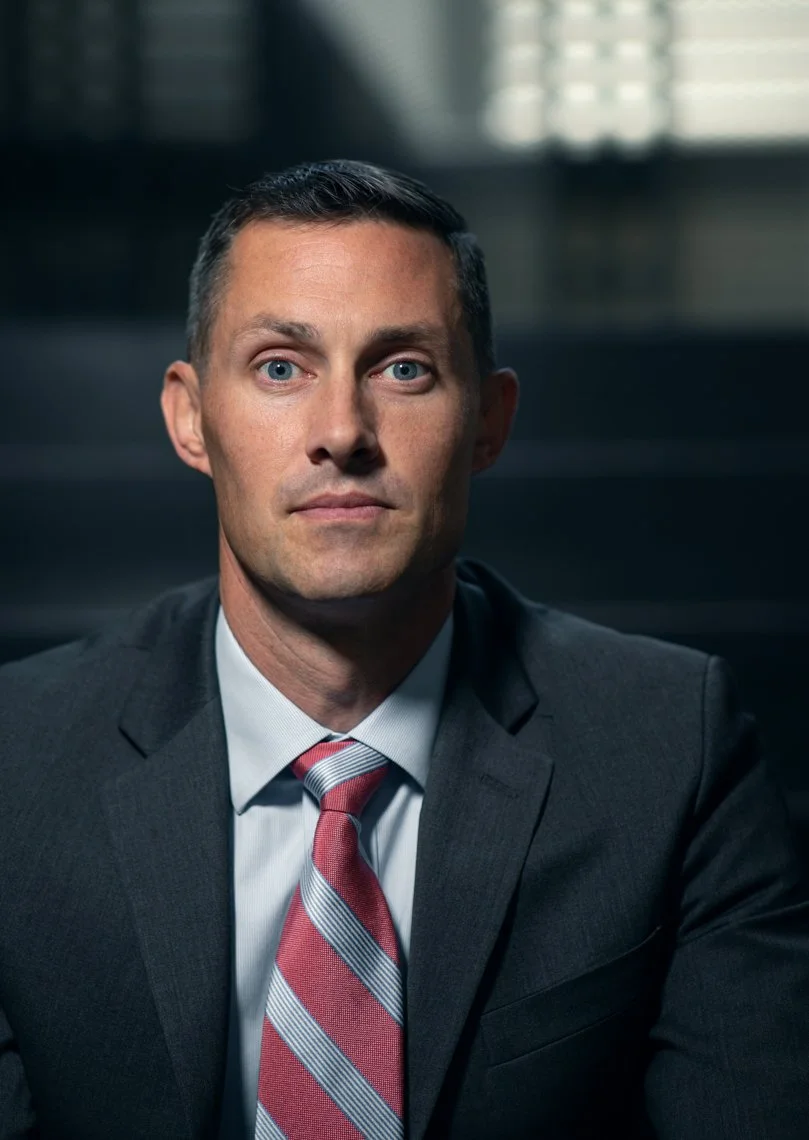 A man in a dark suit, white shirt, and striped red and gray tie sitting in a professional setting with a serious expression.