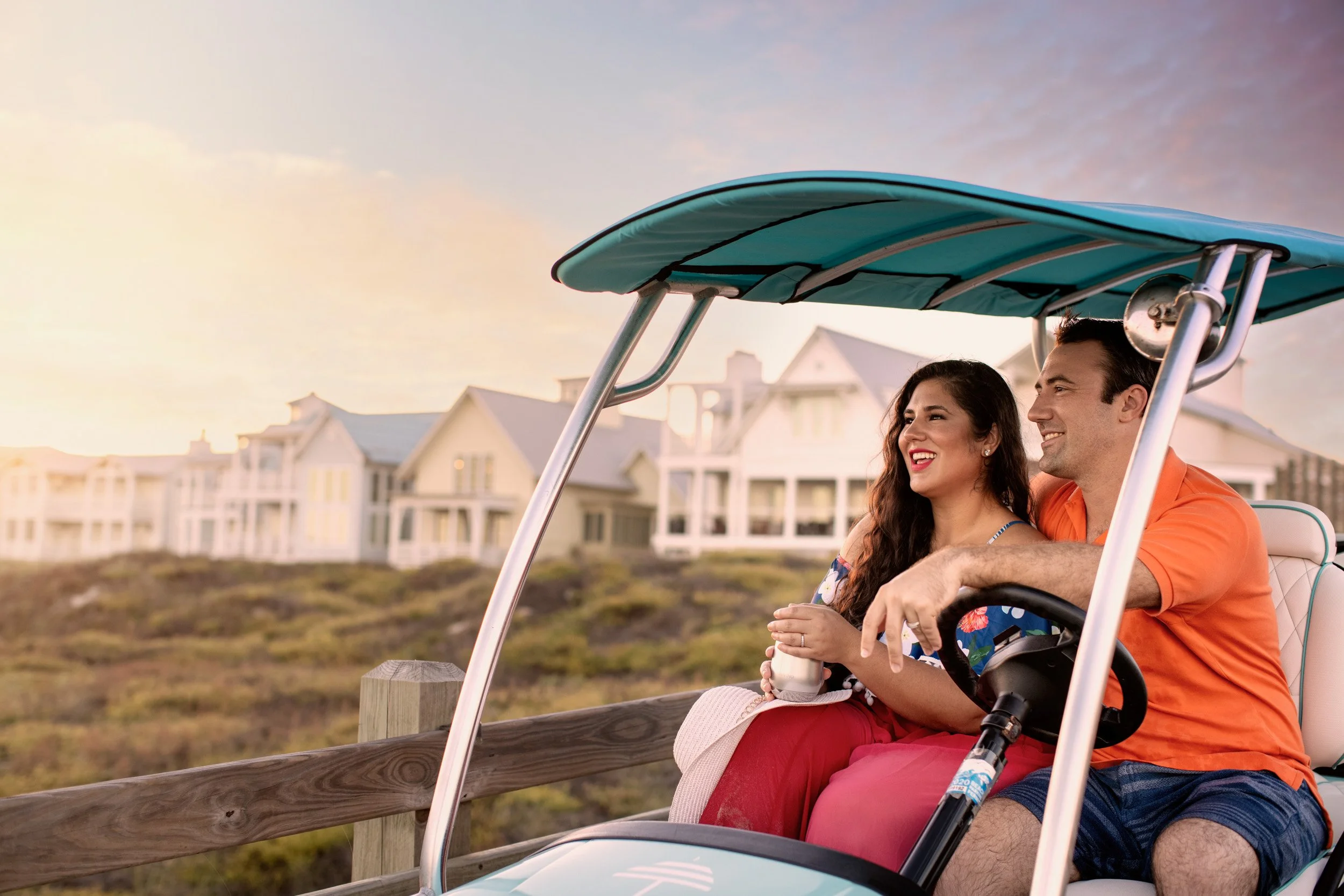 A couple sitting on a golf cart, enjoying a sunset with white houses in the background.