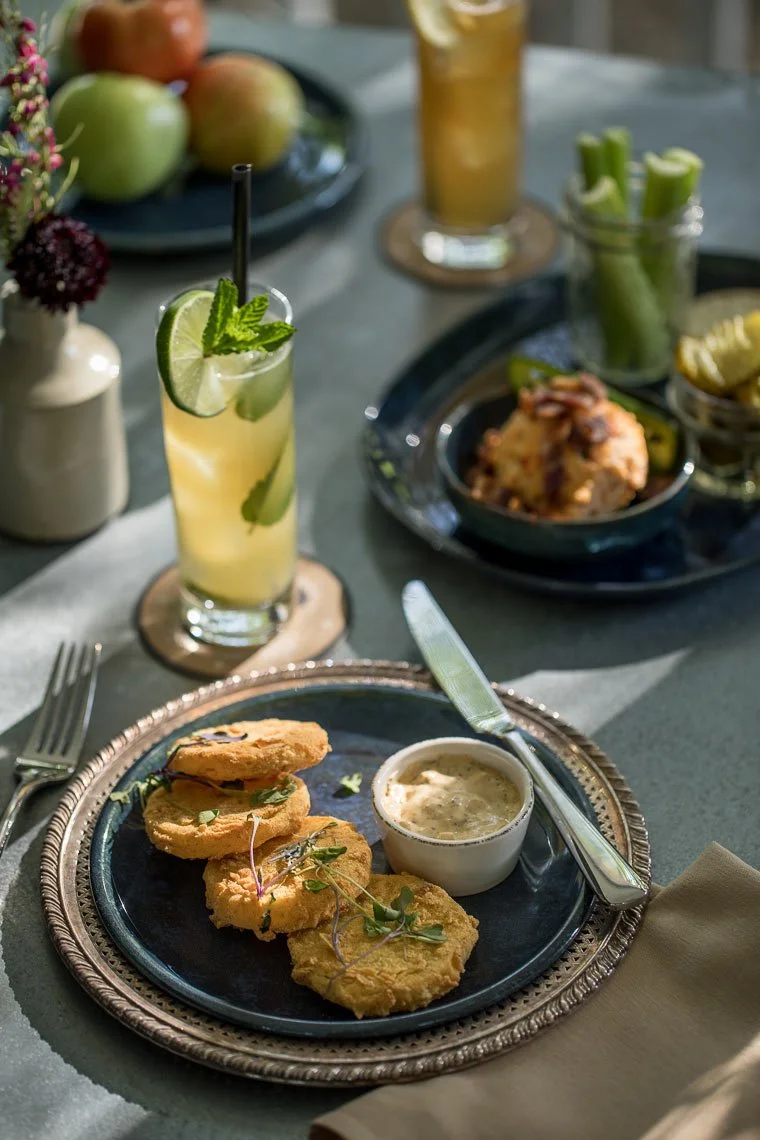 Plate of fried fish with tartar sauce, a decorated drink with lime and mint, and various appetizers and drinks on a dinner table setting.