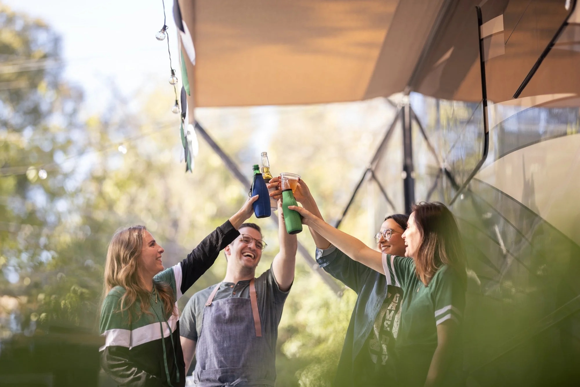 A group of friends cheer and clink bottles in an outdoor setting under a canopy, toasting to good times.