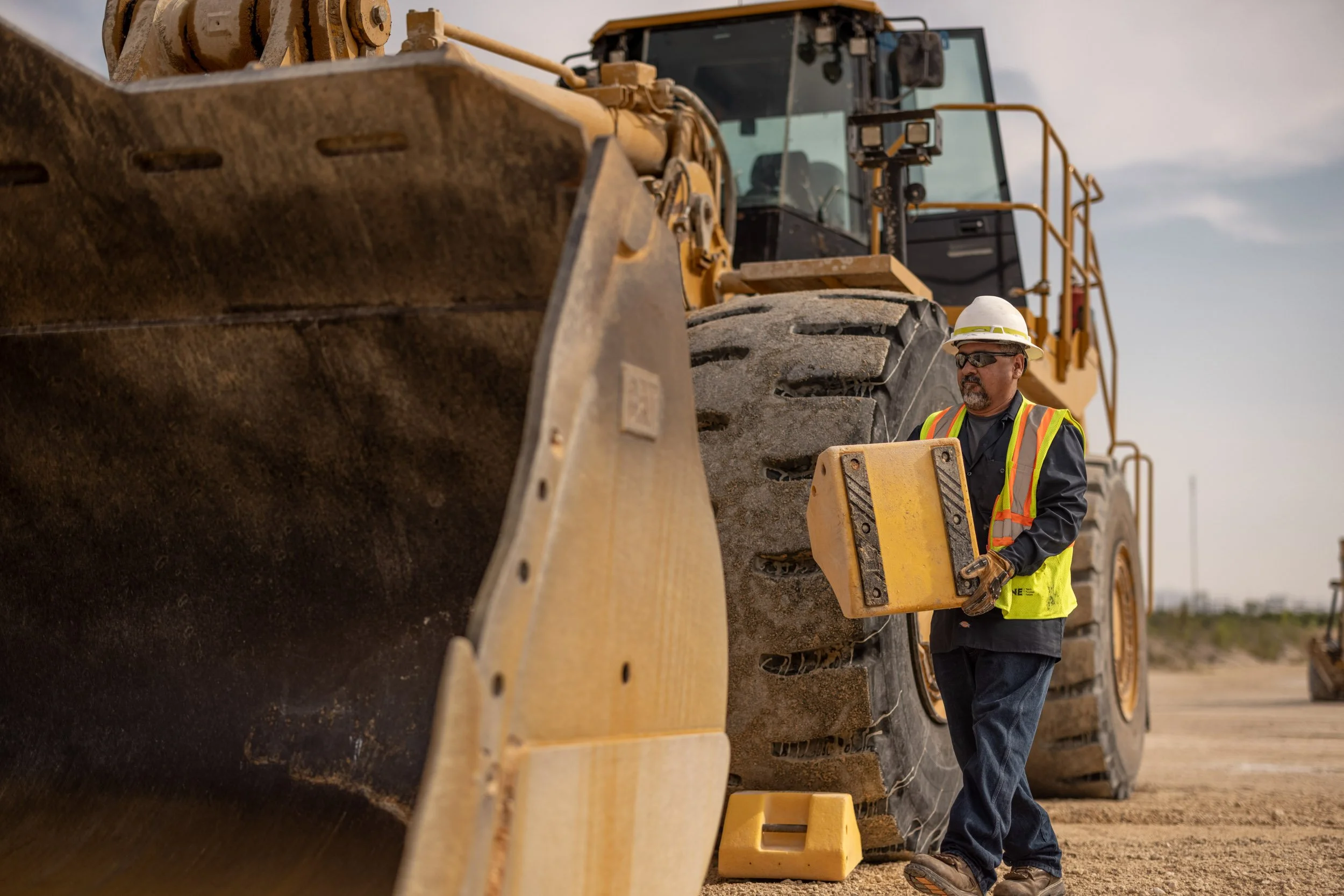 A construction worker wearing a hard hat, safety glasses, and a reflective safety vest supervises a large yellow bulldozer on a dirt construction site.