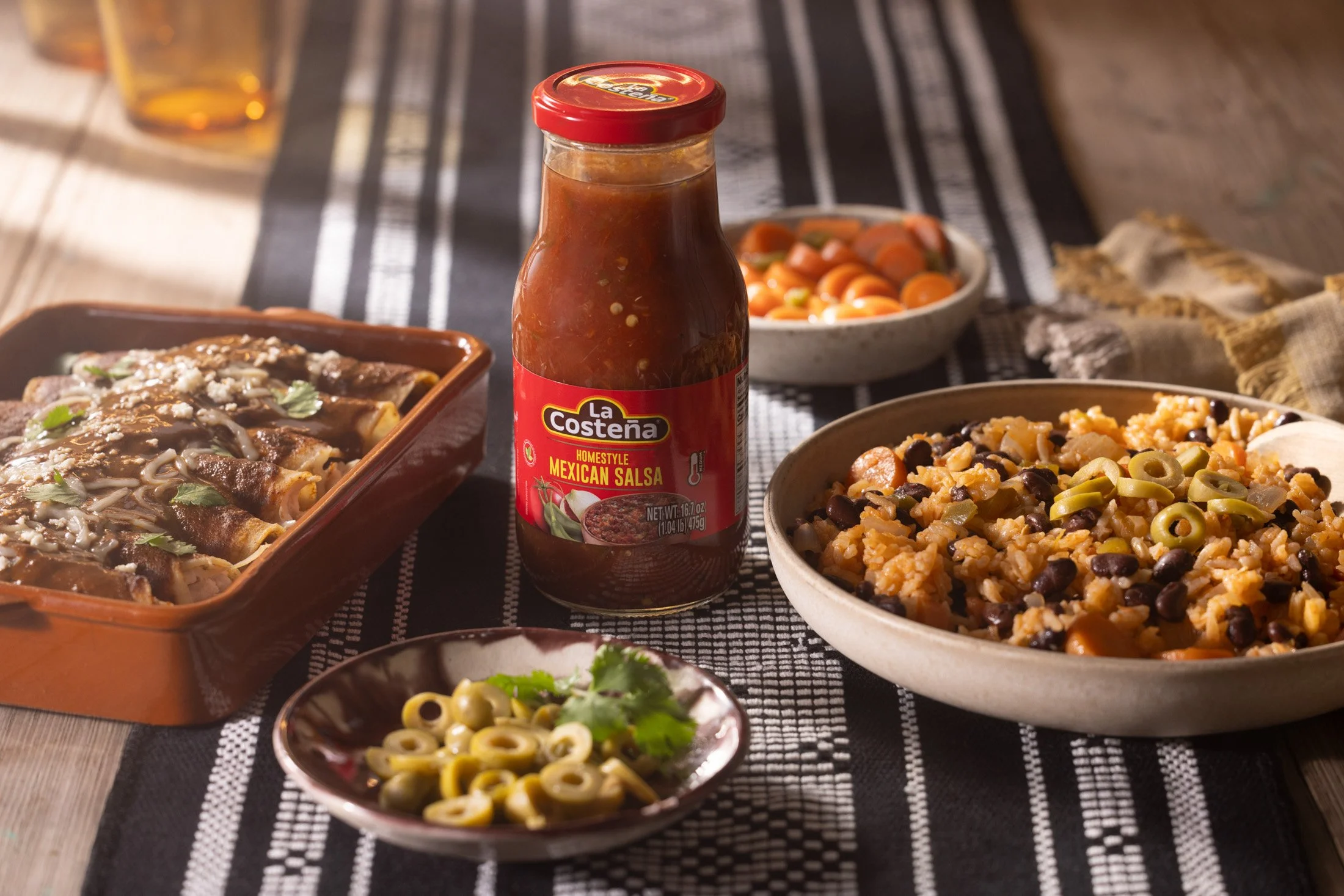 Table with various Mexican food dishes including pasta, salad, and salsa, with a bottle of La Costeña Mexican salsa in the center.