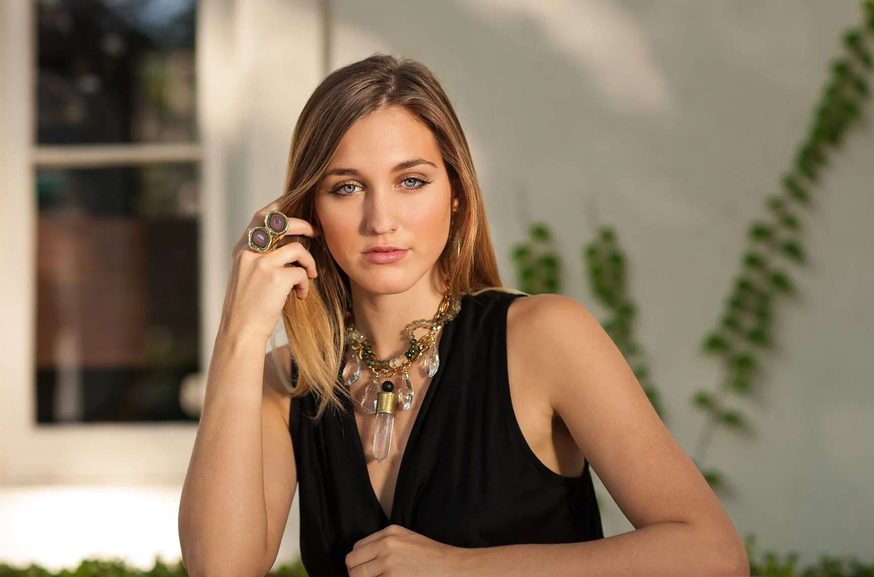A woman with light brown hair and blue eyes wearing a black sleeveless top, multiple necklaces, and rings, sitting indoors near a window with green plants in the background.