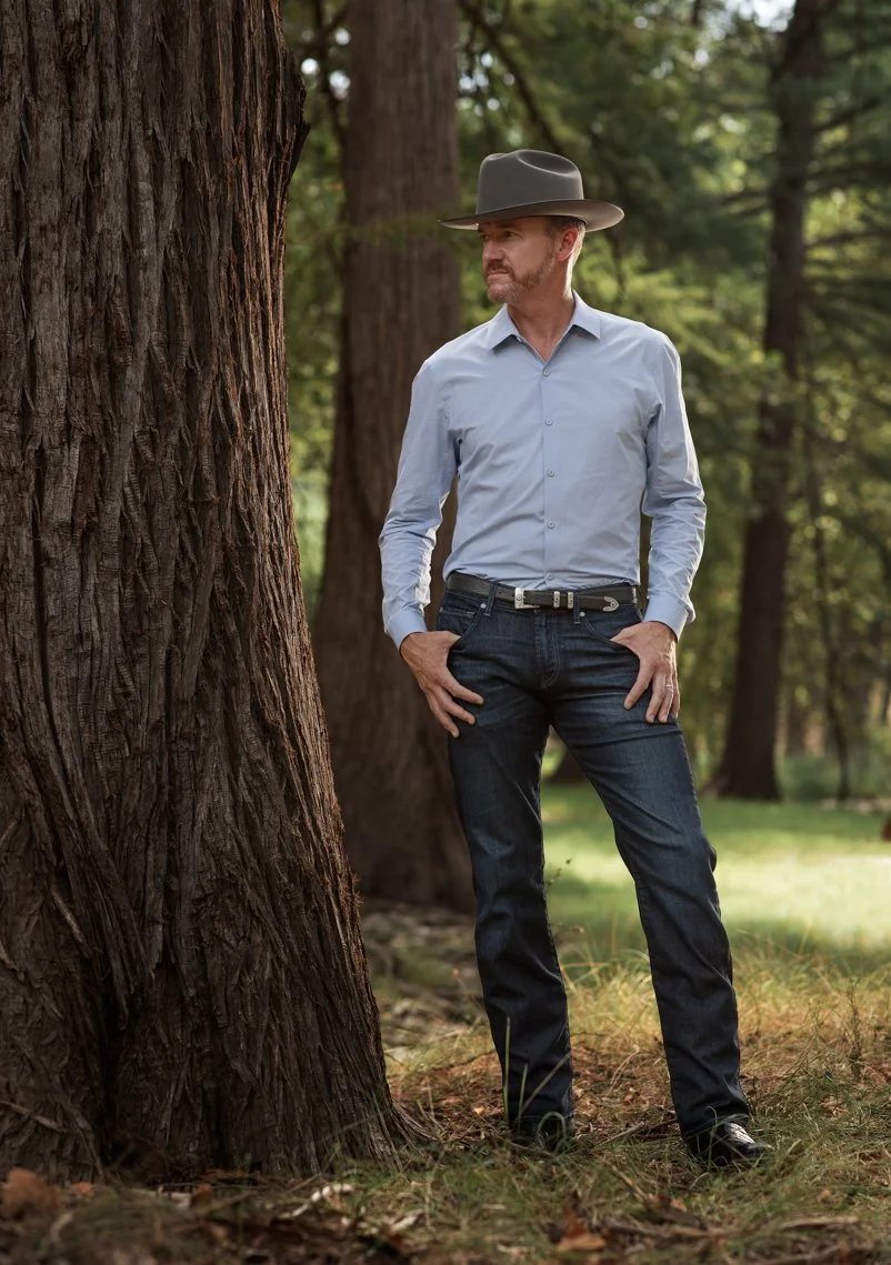 A man in a light blue dress shirt, dark jeans, and a wide-brimmed hat standing outdoors in a forest, with tall trees in the background and one hand on his hip.