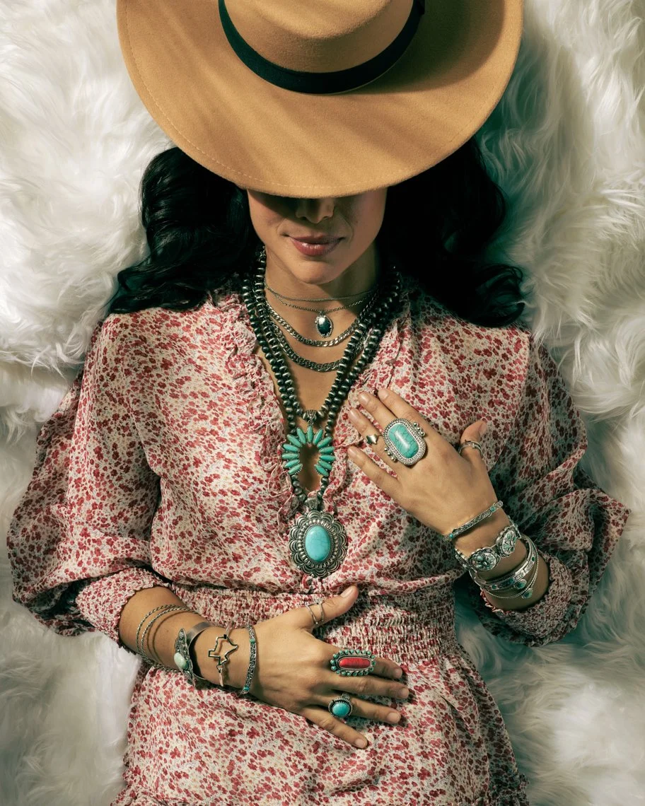 A woman lying on a fluffy white surface, wearing multiple layers of turquoise jewelry, a pink and white patterned dress, and a wide-brimmed hat that covers her eyes.