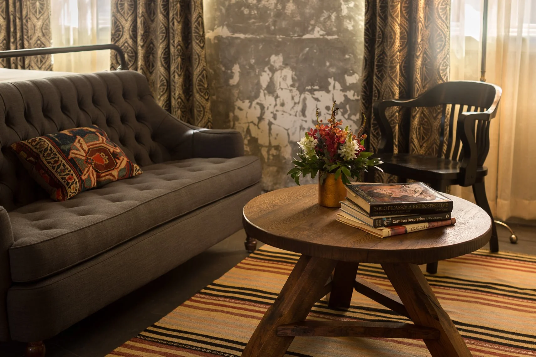Living room with a gray sofa, black wooden chair, a round wooden coffee table with a stack of books and a flower arrangement, patterned curtains, and a striped rug.
