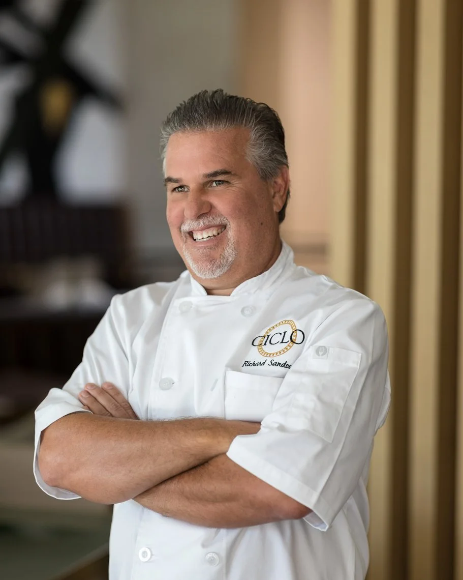 A smiling middle-aged man in a white chef's coat standing with arms crossed in a restaurant kitchen.