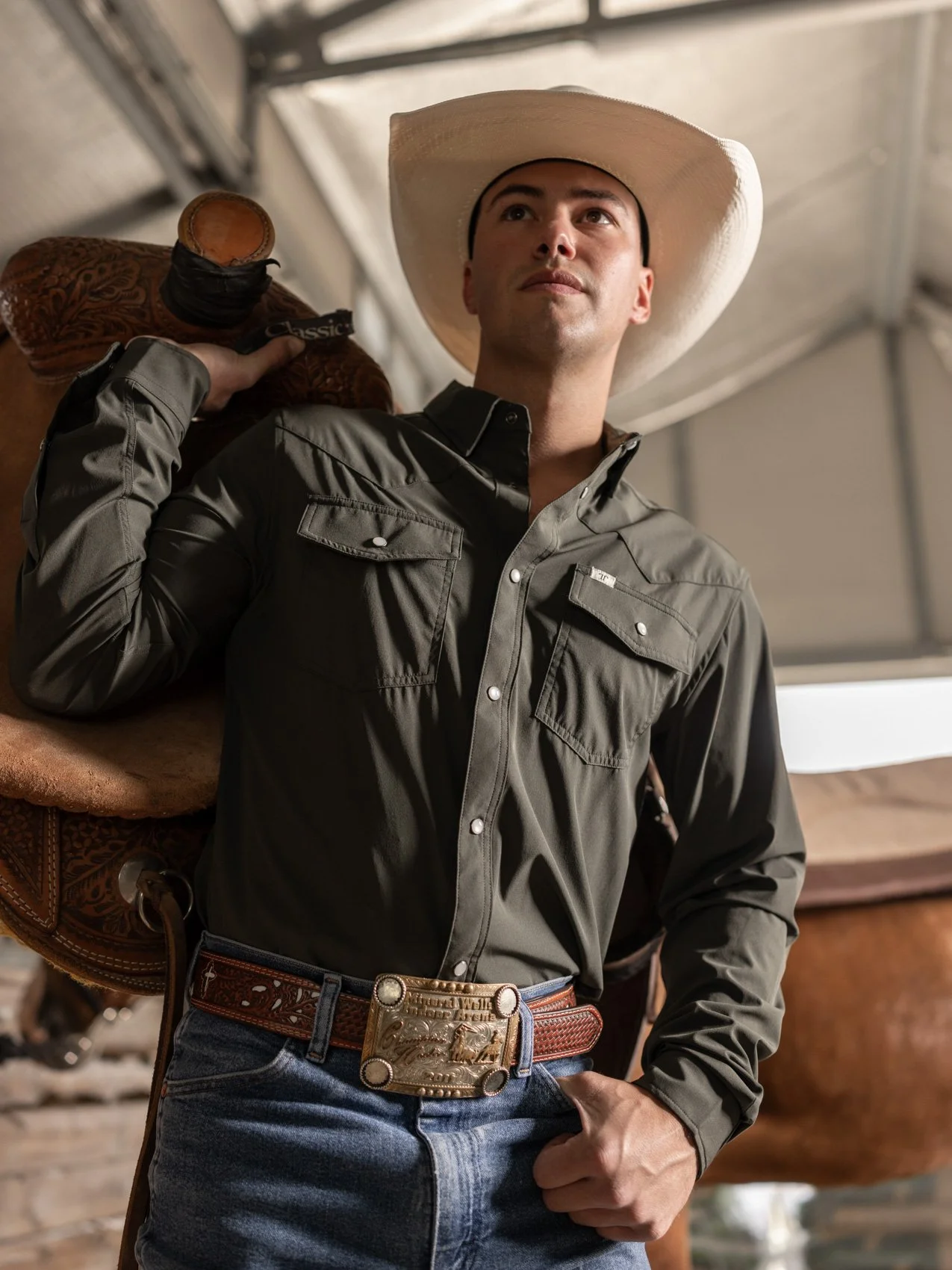 Man wearing a cowboy hat, gray button-up shirt, and jeans, standing with one hand on his belt and the other holding a saddle, inside a barn or stable.