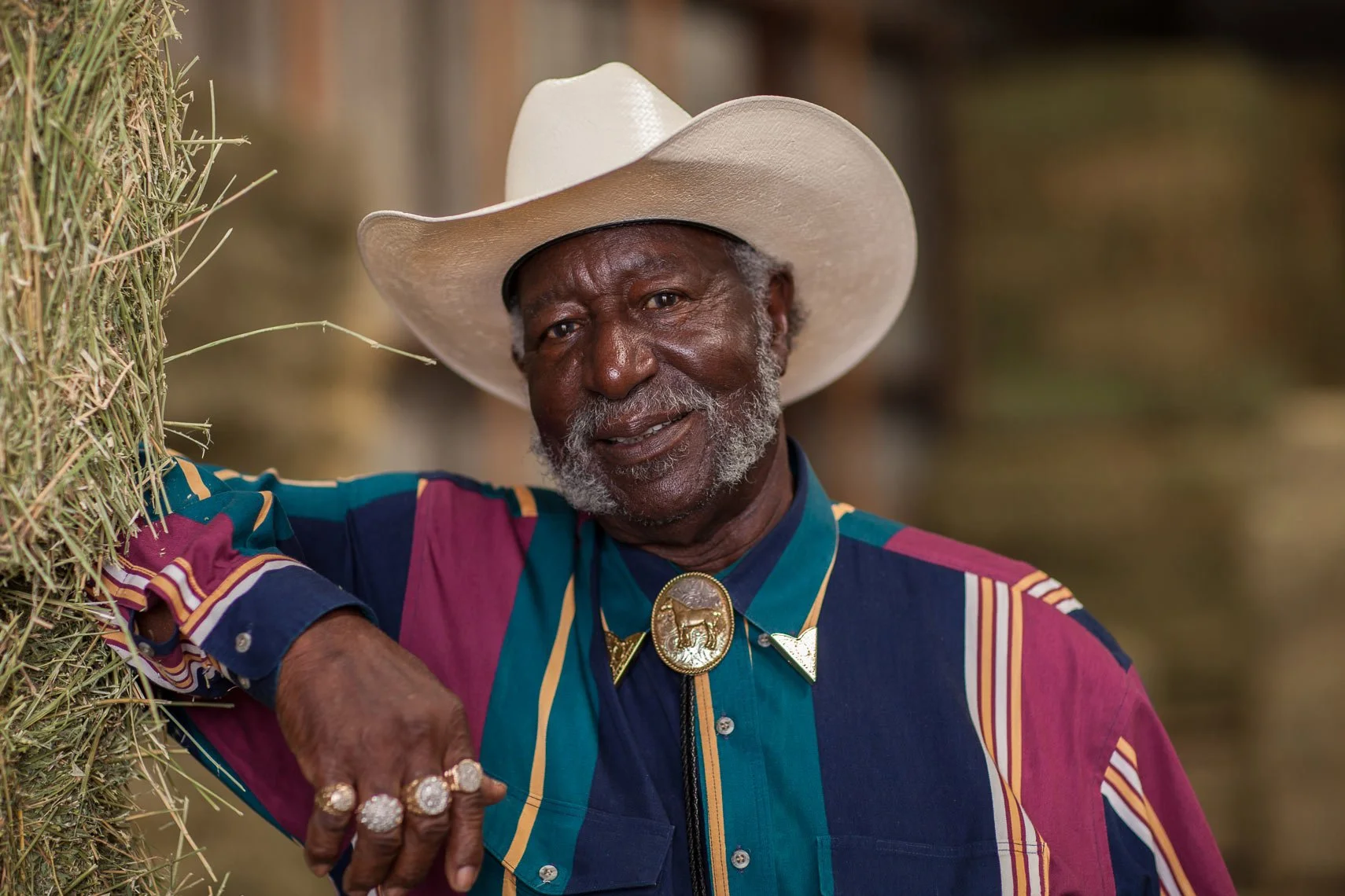 An elderly African American man wearing a wide-brimmed white cowboy hat and colorful striped shirt, standing outdoors near a haystack, smiling and showing off multiple rings and a decorative bolo tie.