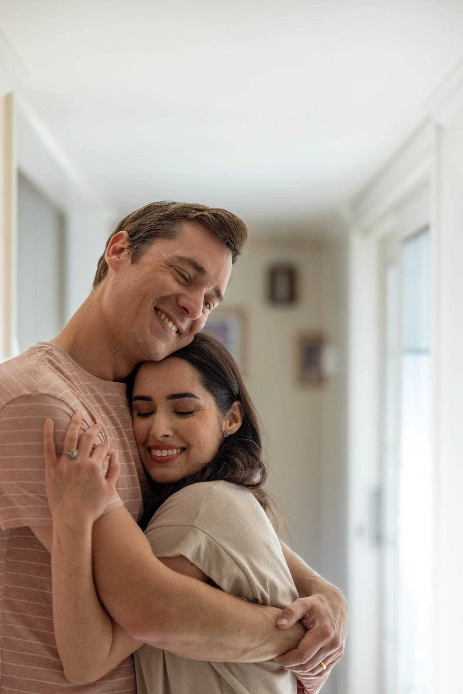 A couple hugging and smiling in a bright room, with the man slightly taller and resting his head on the woman's head, both looking happy and content.